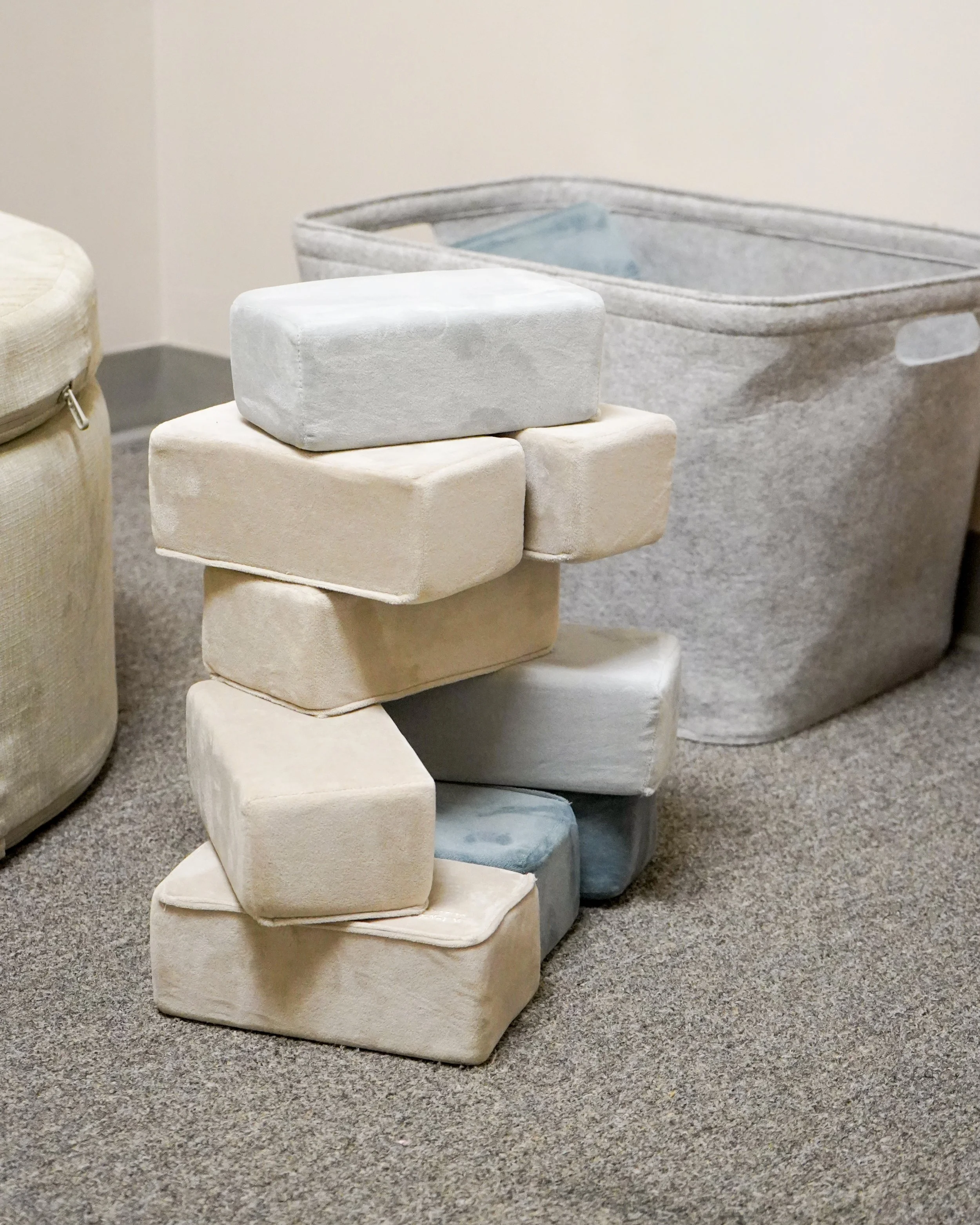 Stacked soft blocks or cushions in pastel shades beside a storage container on a carpeted floor.
