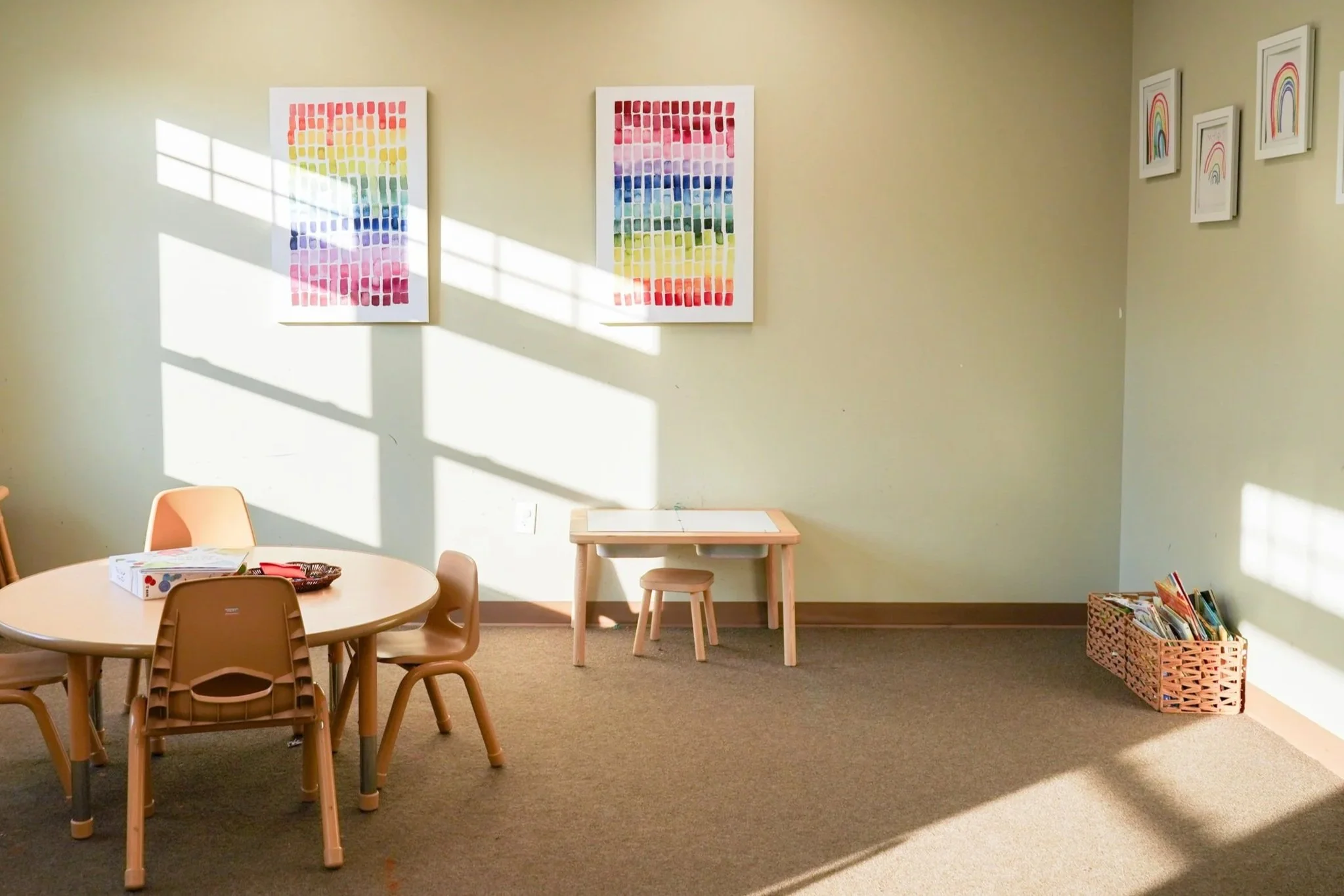 A room with educational artwork on the walls, a round table with chairs, and a basket of books on the floor, with sunlight casting shadows through a window.