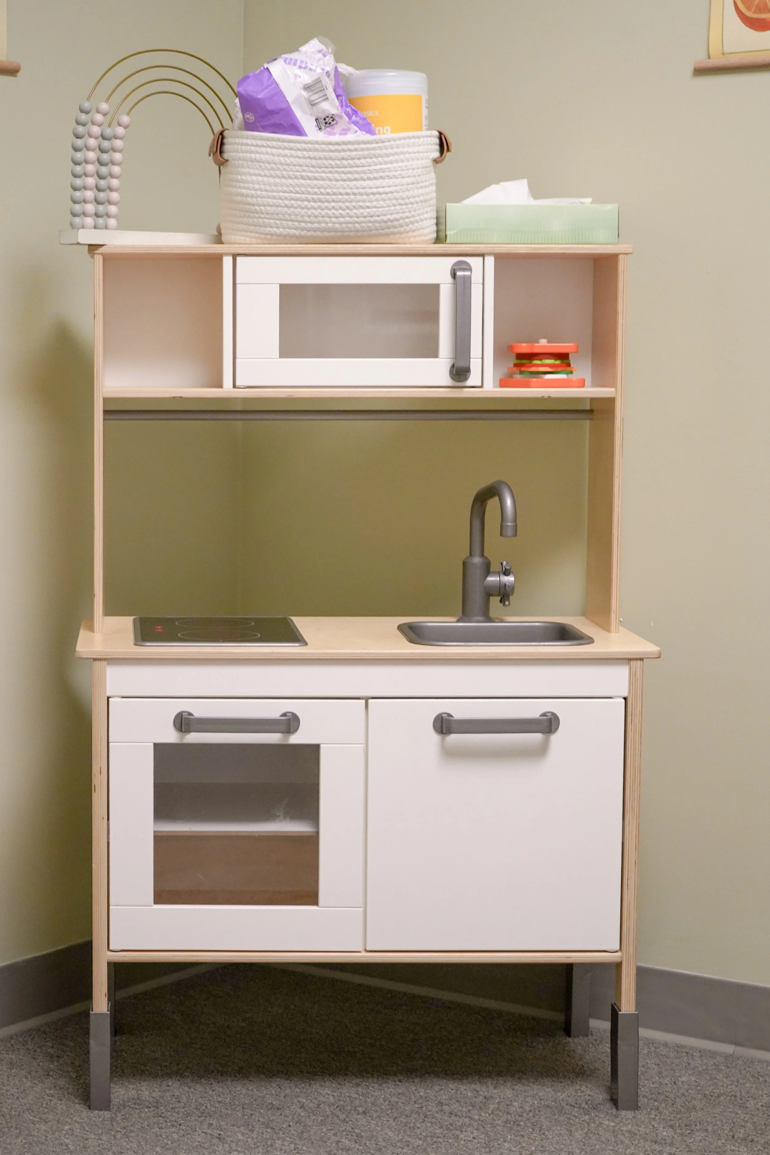 A small wooden play kitchen with white doors and a black cooktop, featuring a sink with a silver faucet, set against a beige wall.