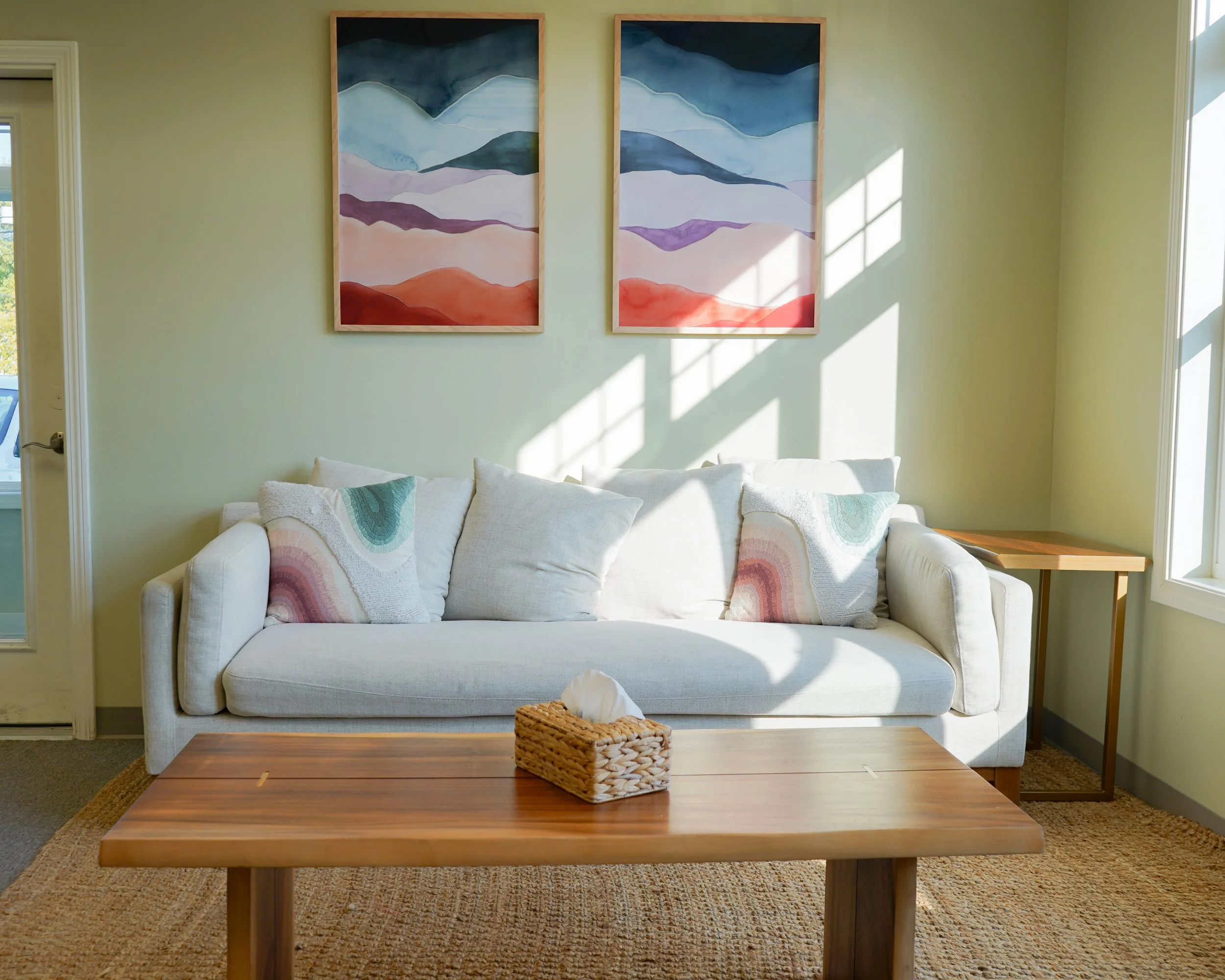 Living room with a white sofa, colorful throw pillows, wooden coffee table with tissue box, wall art of abstract mountain landscape, a side table, and sunlight streaming through a window.