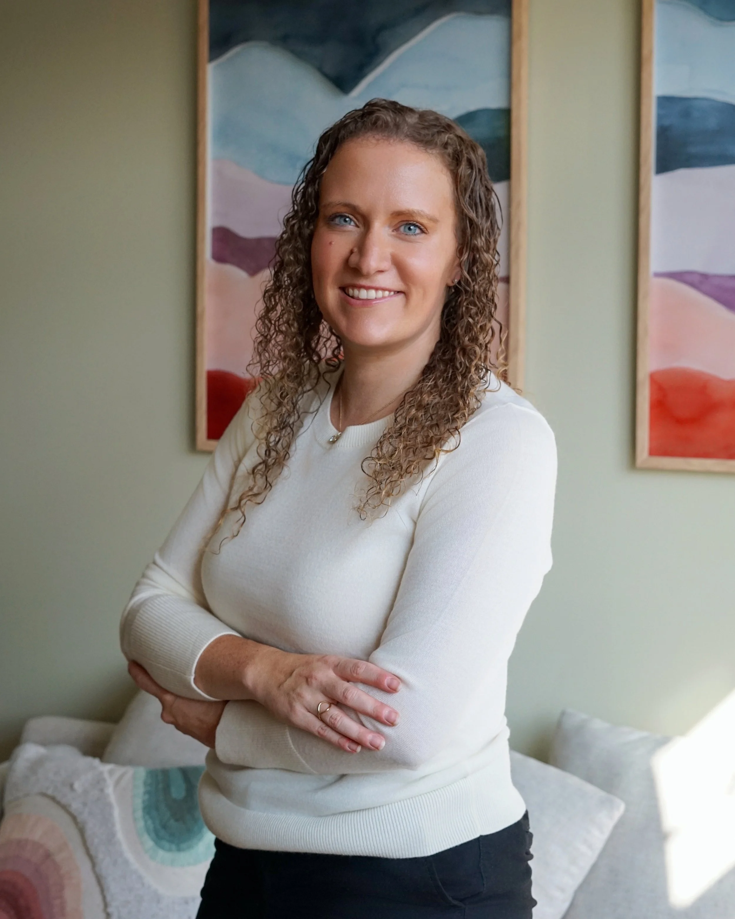 A woman with curly hair and blue eyes standing with arms crossed in a cozy living room, with colorful abstract paintings on the wall behind her.