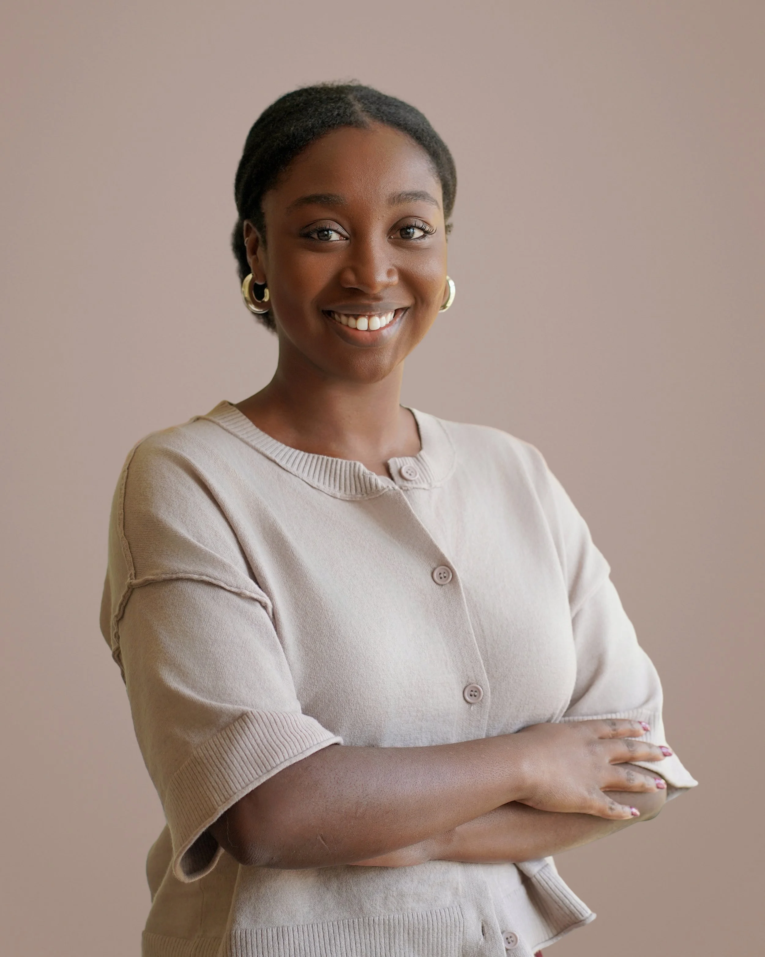 A woman with short, dark hair wearing a beige cardigan, smiling and standing with arms crossed in front of a neutral background.