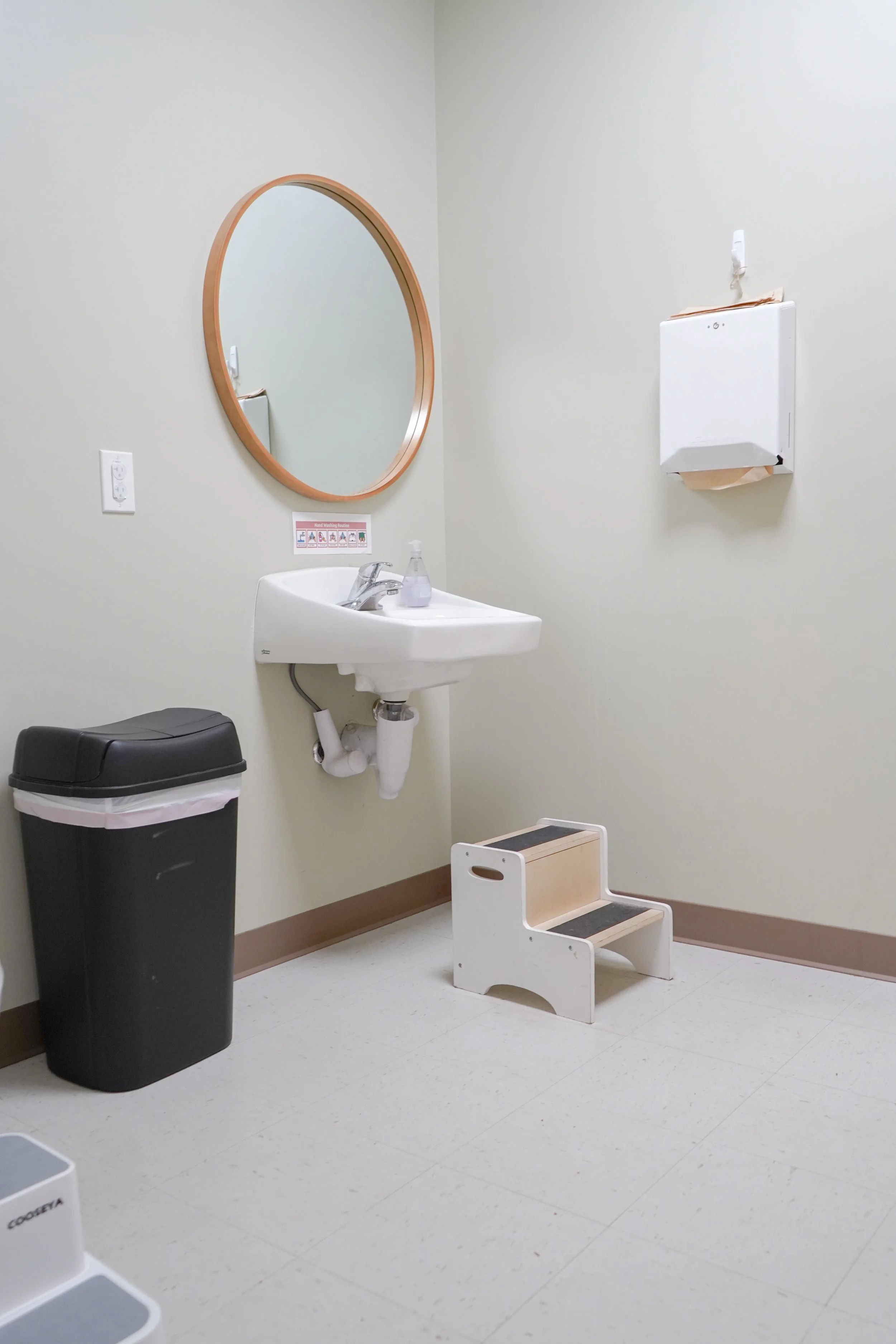 A restroom with a small sink, a mirror, a step stool, a trash can, a paper towel dispenser, and a soap dispenser on the sink.