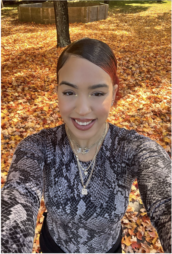 A woman with dark hair and makeup smiling and taking a selfie outdoors on a fall day with colorful fallen leaves and a tree in the background.