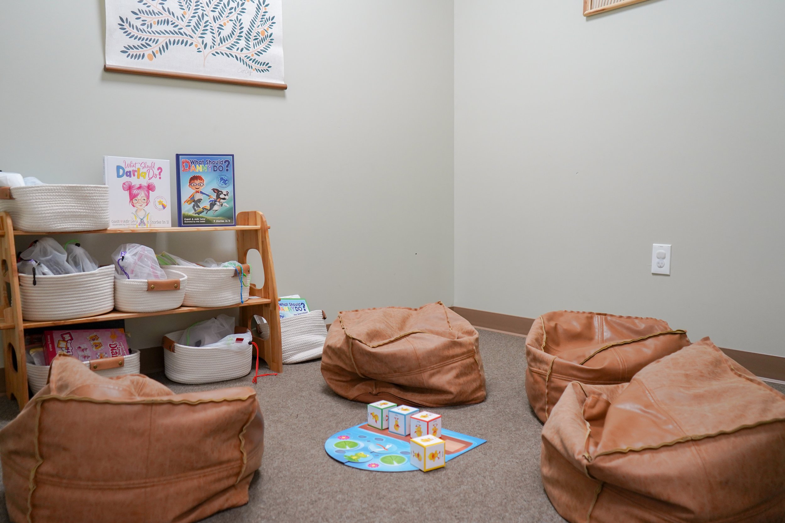 Children's reading and play area with bean bags, a small game board with dice, books on a shelf, and a framed art piece on the wall.