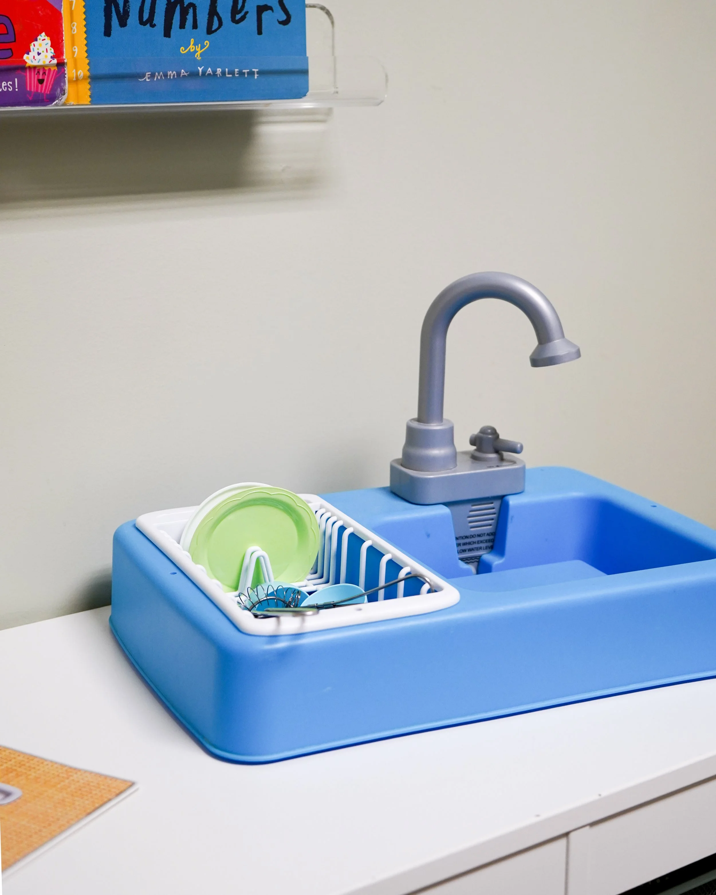 A blue toy kitchen sink with a silver faucet, a white dish rack holding green plates and blue utensils, set on a white countertop in a pretend play area.