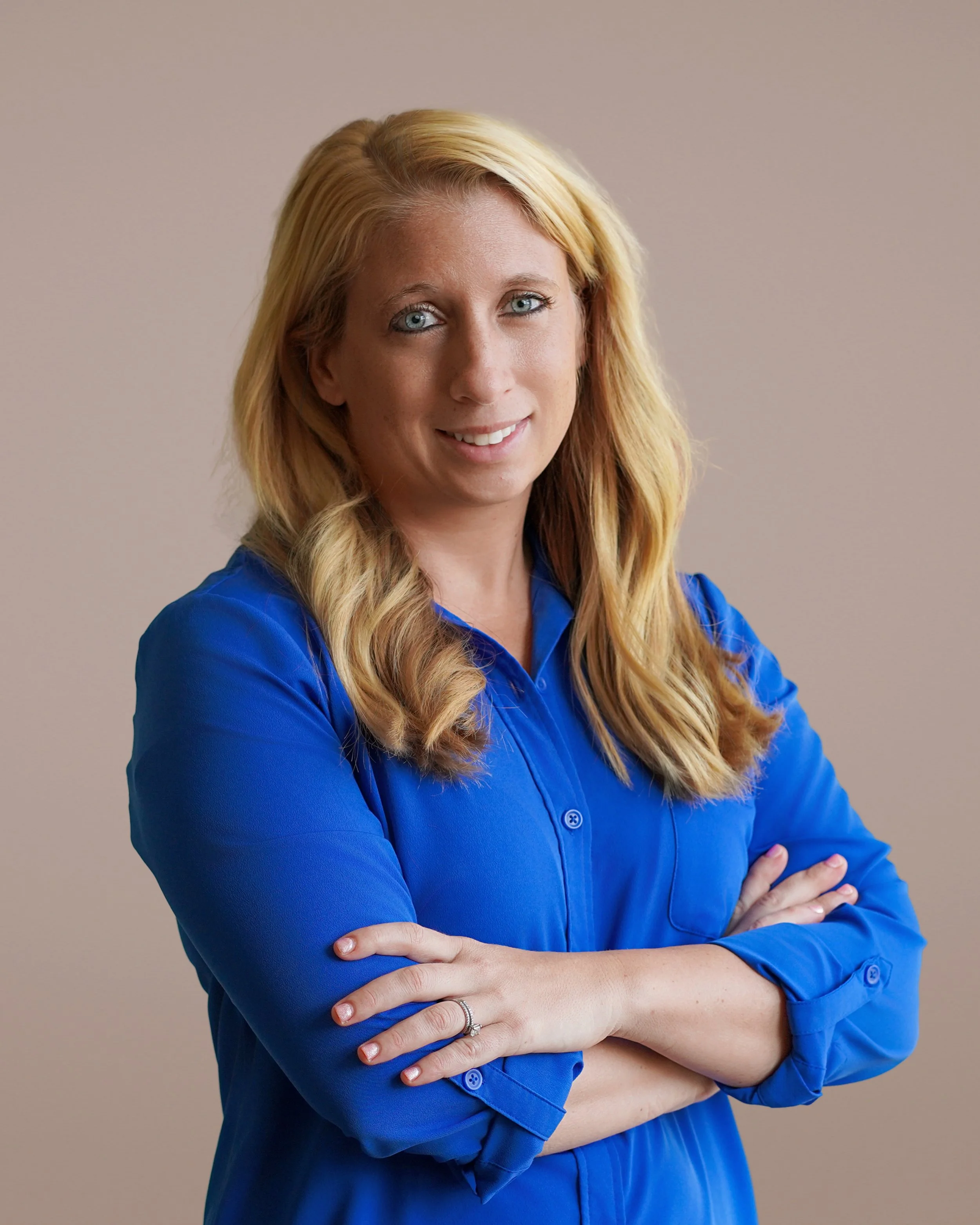 A woman with blonde hair and blue eyes, wearing a blue long-sleeve blouse, standing with arms crossed against a beige background.