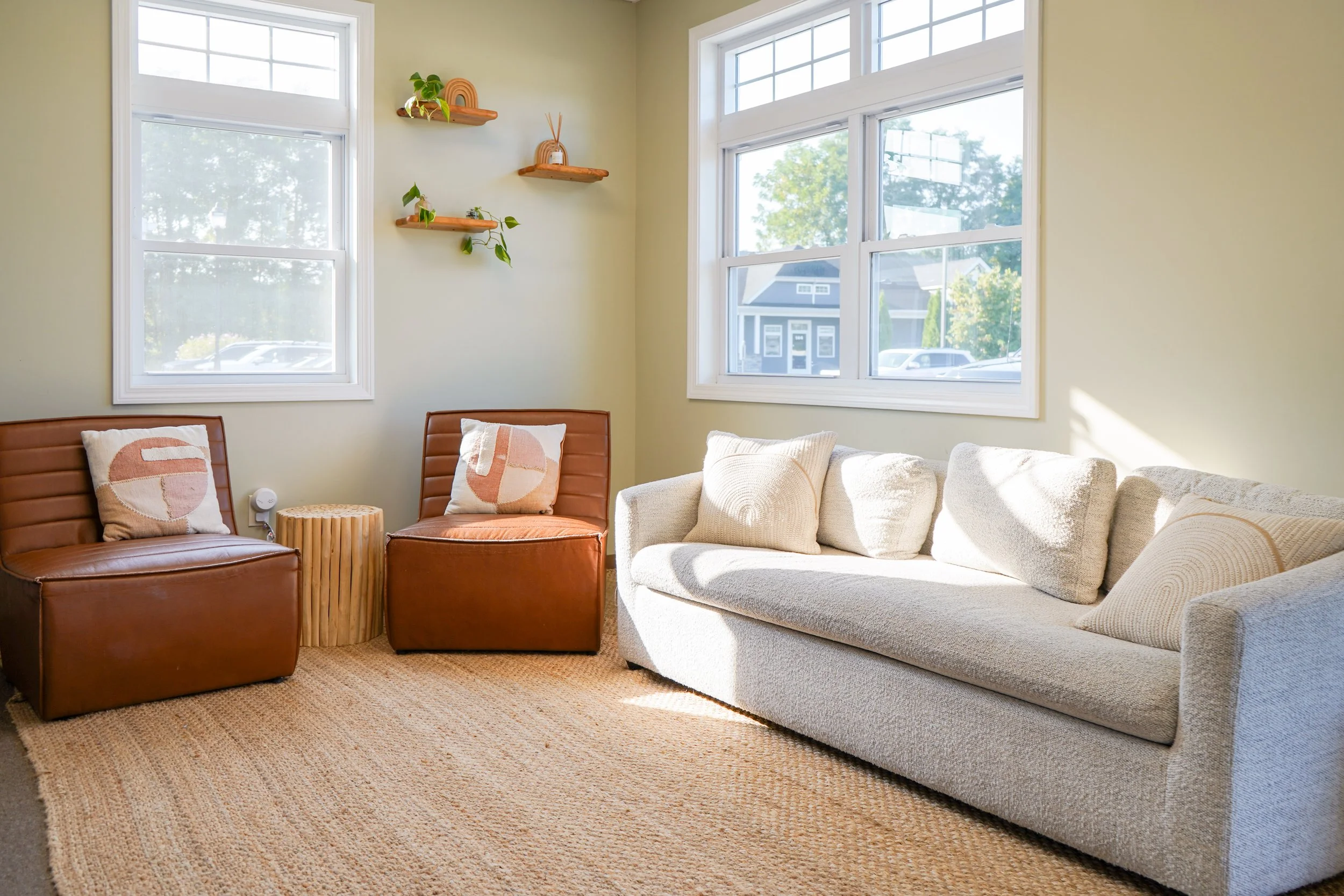 Sunlit living room with two brown leather chairs with decorative pillows, a cream sofa with pillows, a woven rug, and wall-mounted shelves with plants.