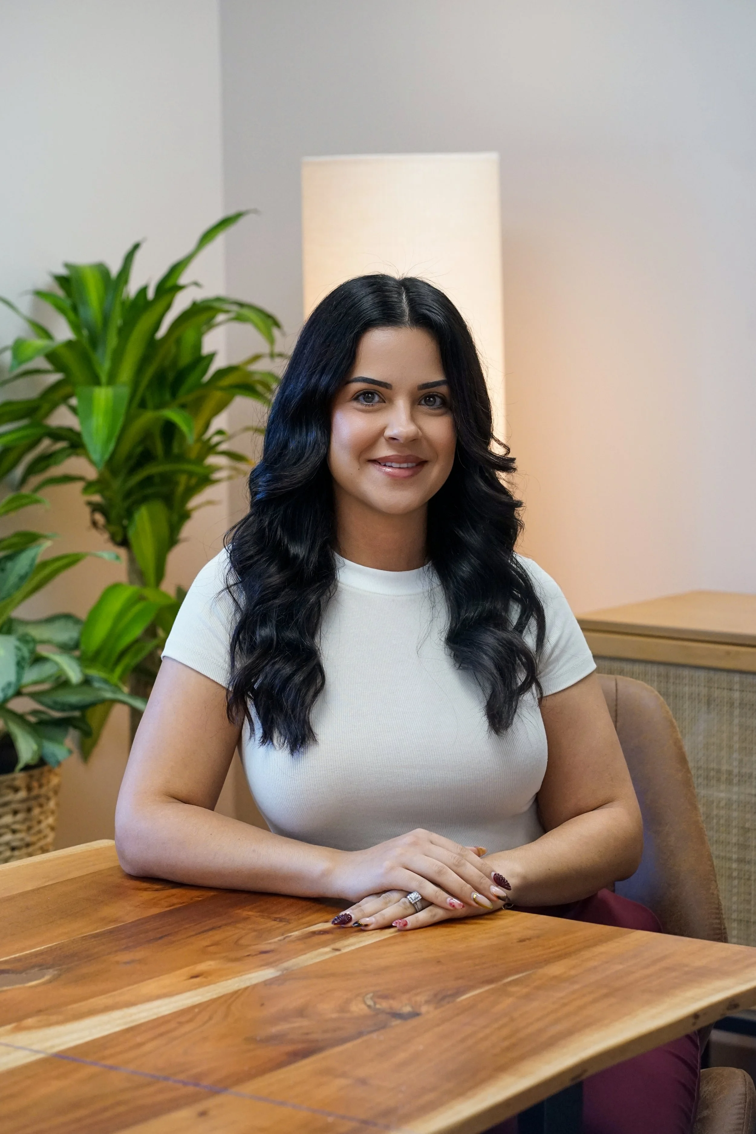 A woman with long black wavy hair, wearing a white t-shirt, sitting at a wooden table in a room with a potted plant and a lamp in the background.