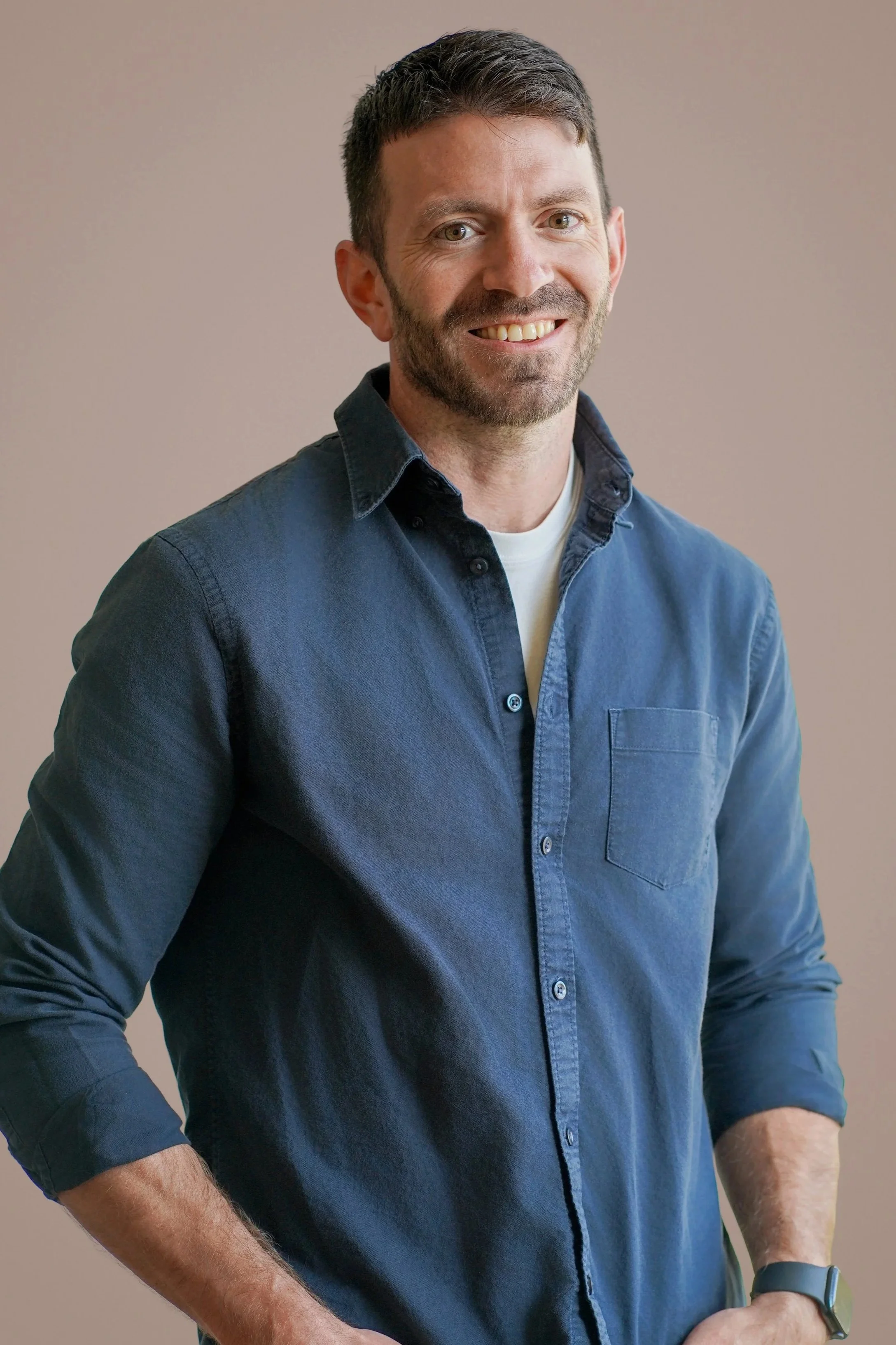 A smiling man with short dark hair and a beard, wearing a dark blue button-up shirt and a watch on his left wrist, standing against a plain beige background.