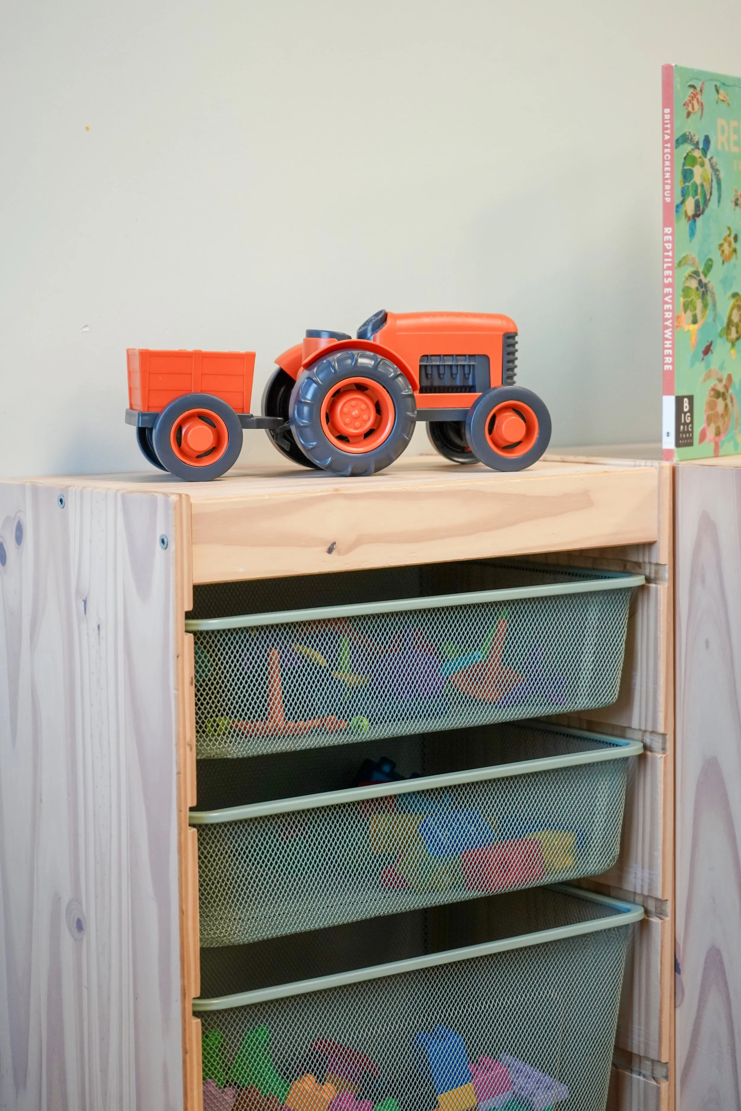 A toy orange tractor with a trailer on top of a wooden storage unit with mesh drawers filled with various toys and a children's book next to it.