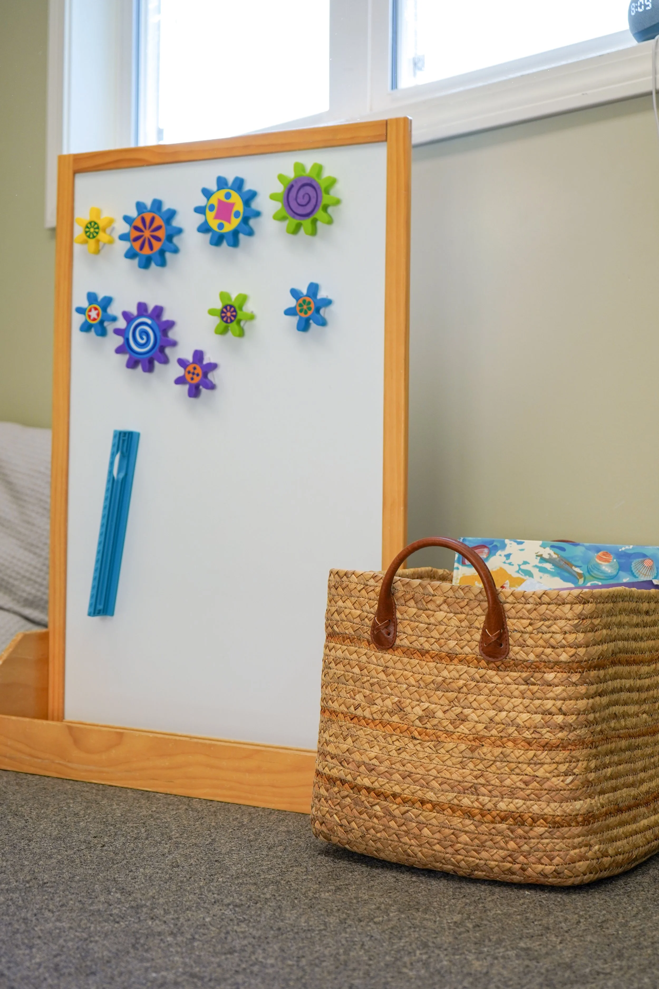 Colorful magnets on a whiteboard with a blue magnetic strip, next to a woven basket on a gray carpeted floor near a wall and window.