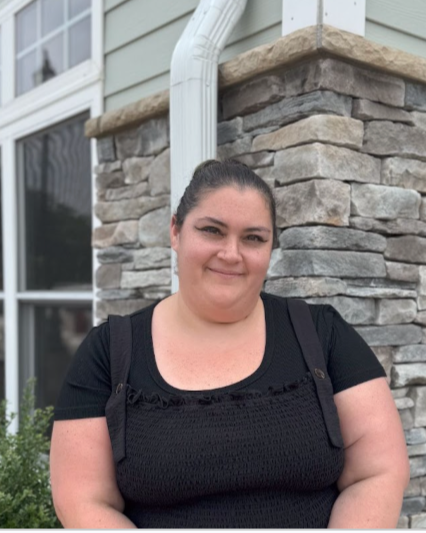 A woman standing outside near a house with stone siding and a window, wearing a black shirt and carrying a backpack.