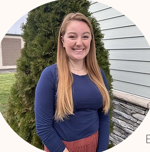 A smiling young woman with long, straight blonde hair standing outdoors near a tree and a house with white and stone siding.