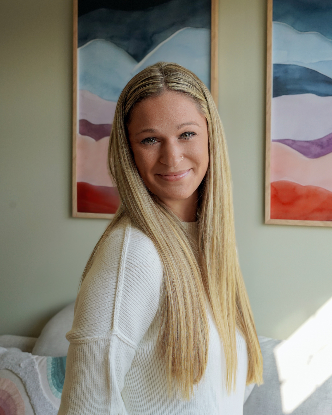 A smiling young woman with long blonde hair, wearing a white sweater, standing in front of colorful mountain paintings on a neutral wall.