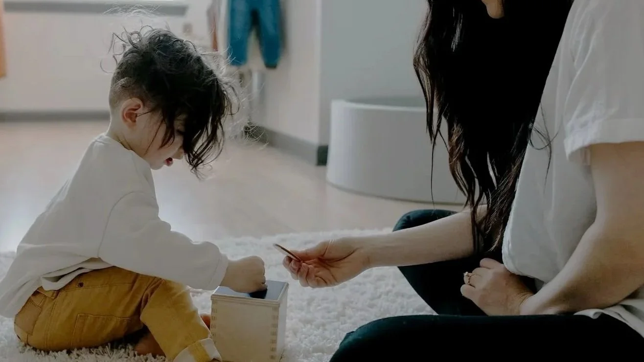 A young child with messy hair sitting on a fluffy rug, playing with a toy inside a box, as an adult woman offers a toy.