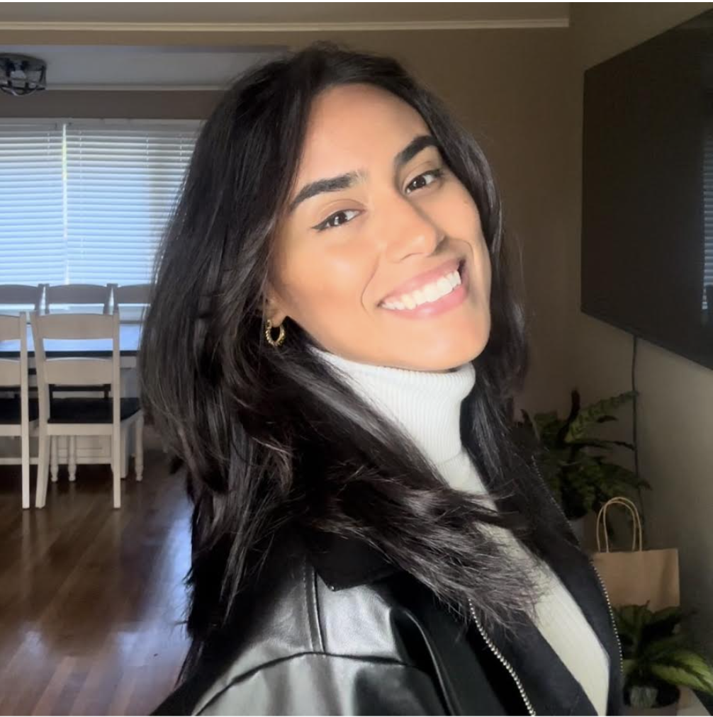 A woman with dark, shoulder-length hair and a bright smile, wearing a white turtleneck and black leather jacket, standing inside a home with a dining area and a large TV in the background.