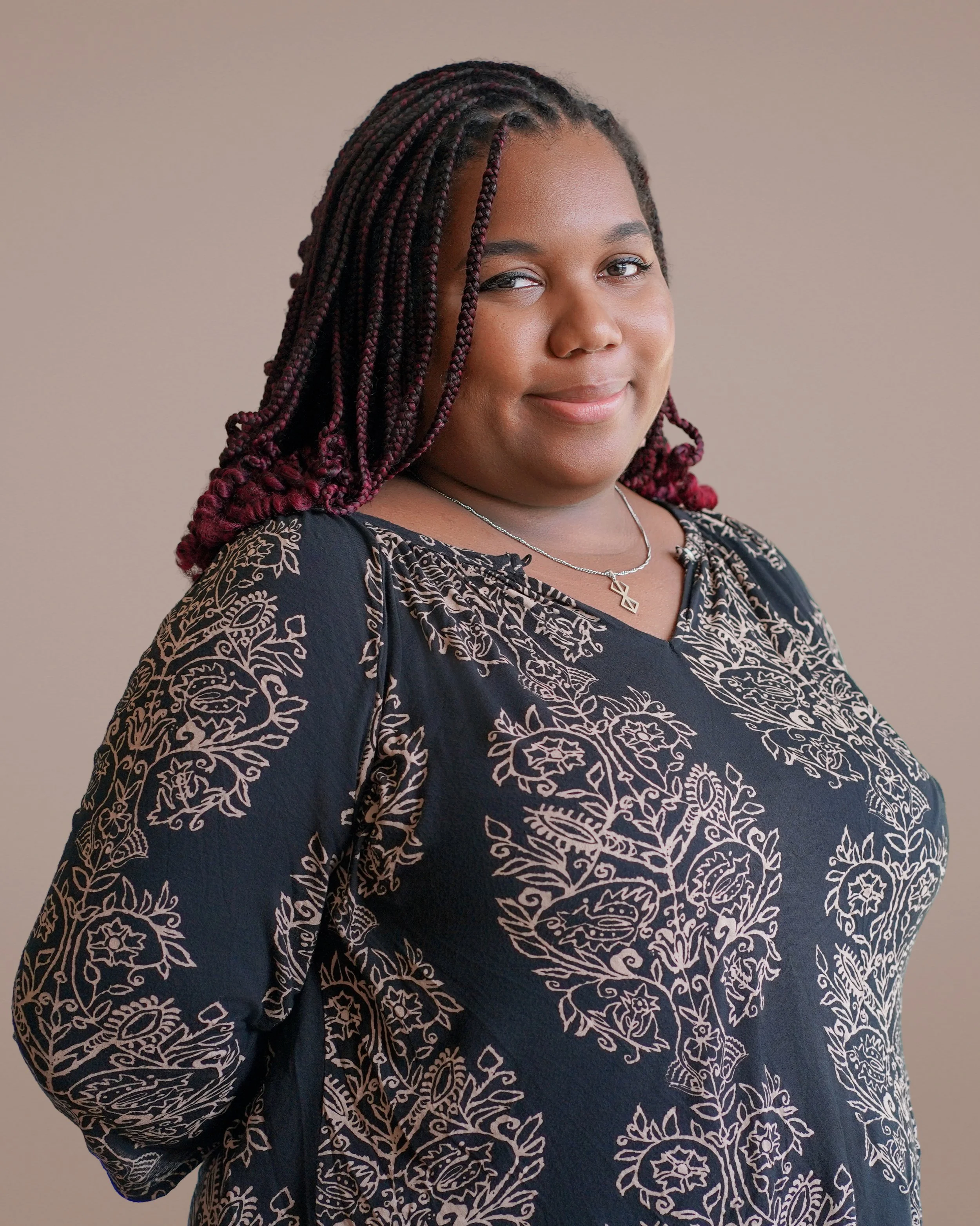 Portrait of a woman with braided hair, wearing a patterned dark blouse and a necklace, smiling softly.