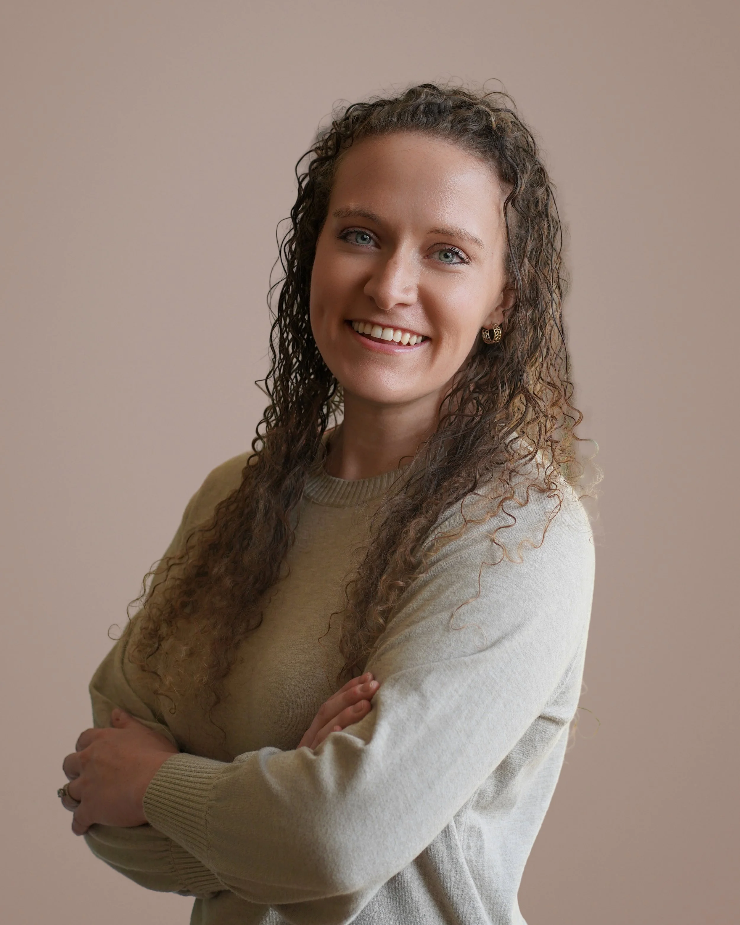 A woman with curly brown hair, blue eyes, and a cheerful smile, wearing a beige sweater and gold earrings, standing with her arms crossed against a neutral background.