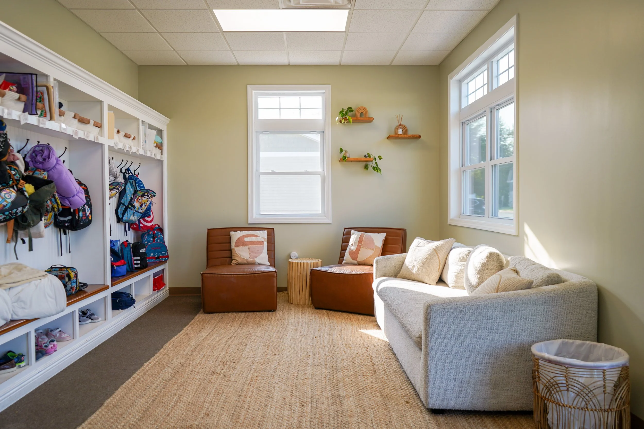 Waiting room with beige walls, a white sofa with beige cushions, two brown chairs with cushions, a small wooden side table, large windows, and shelves with plants, clipboards, and decor.