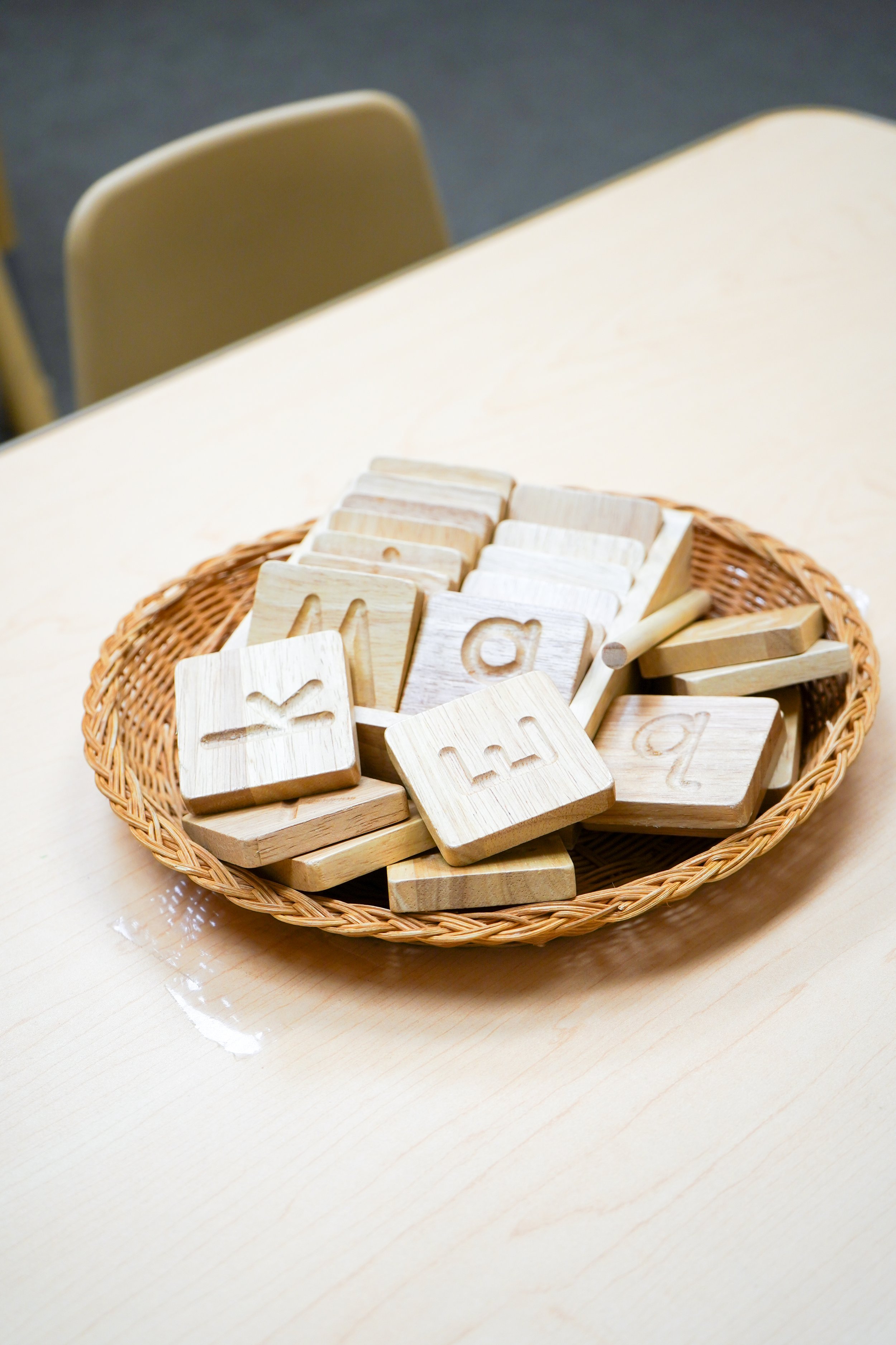Wooden alphabet tiles with carved letters in a wicker basket on a light wooden table.