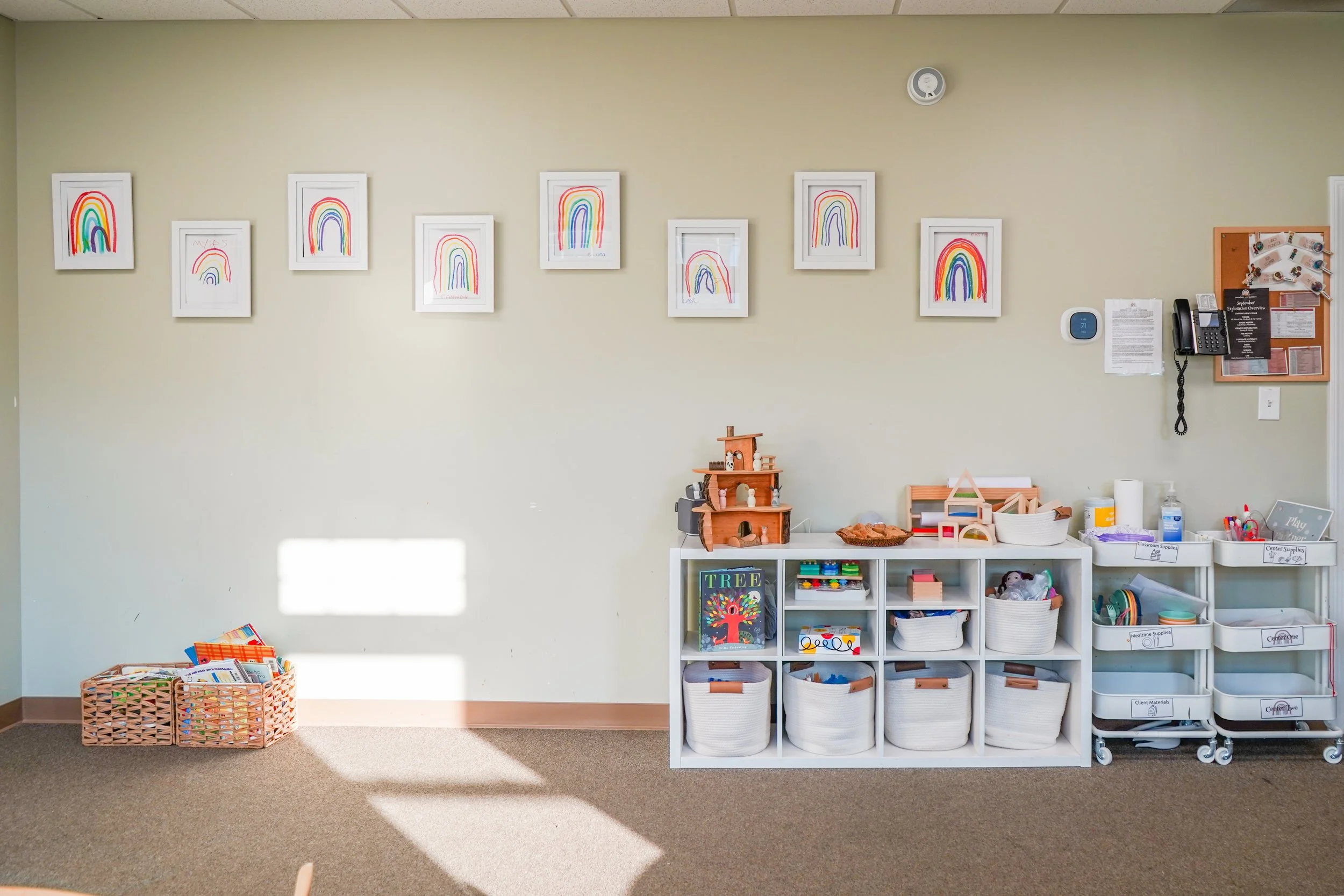 Wall decorated with children’s rainbow drawings, organized toys and supplies on shelves, and a basket of books on the floor in a classroom or playroom.