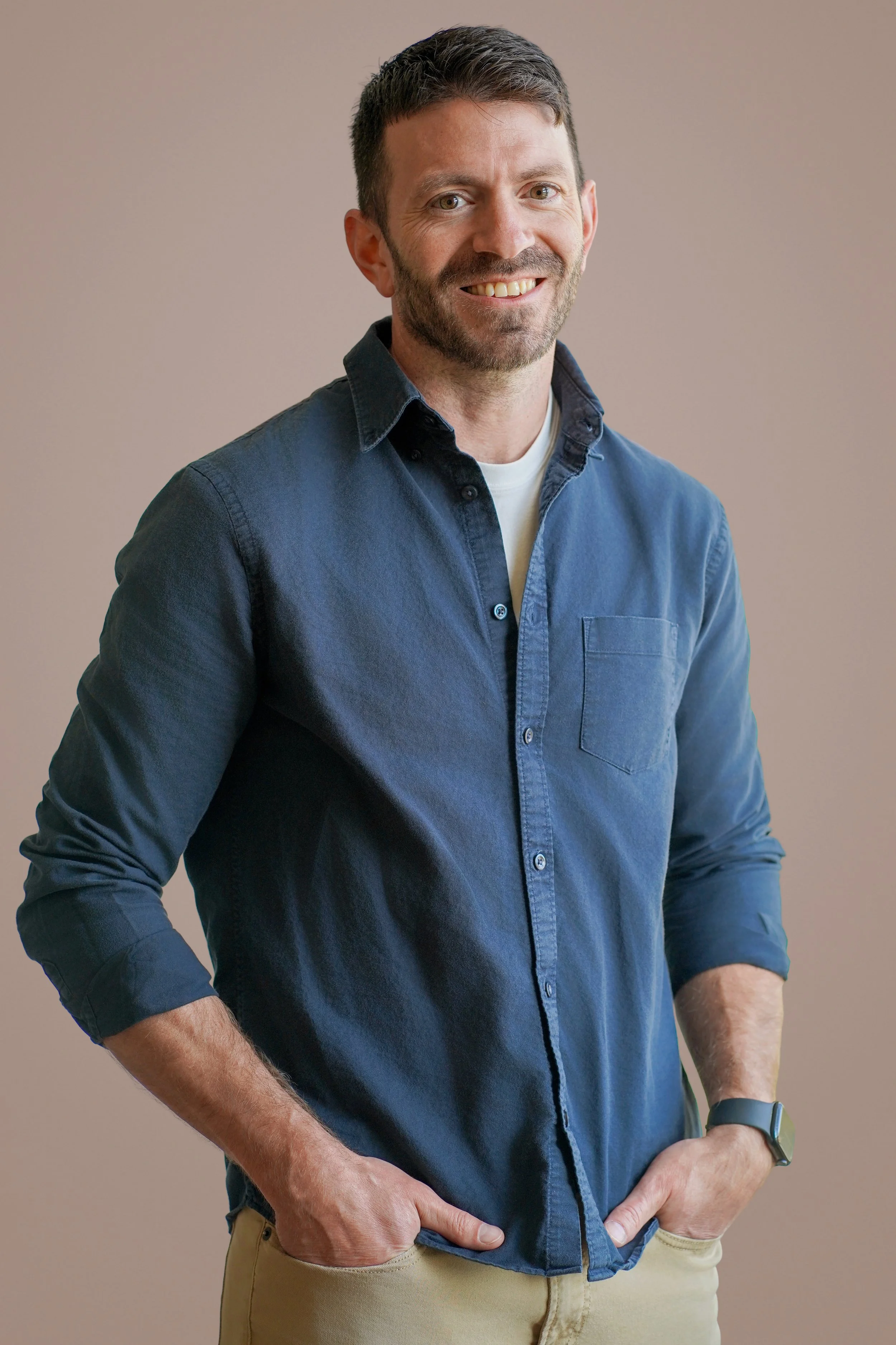 A man with short dark hair, beard, smiling, wearing a navy blue button-up shirt with the sleeves rolled up, standing with hands in pockets against a beige background.