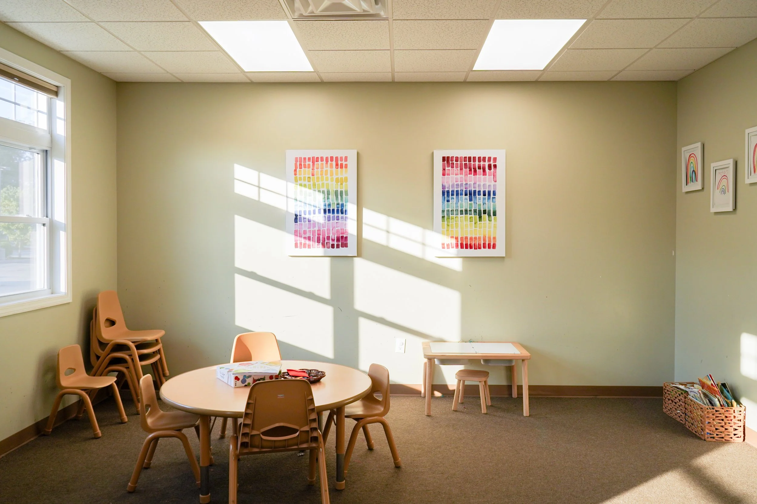 A sunlit children's play room with a round table and six chairs, artwork on the walls, window with blinds, and a basket of books in the corner.