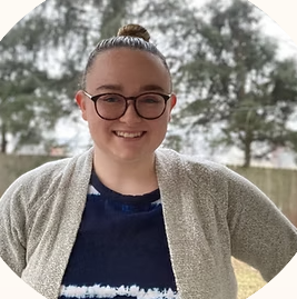 Young woman with glasses smiling outdoors, wearing a gray cardigan and a blue top.