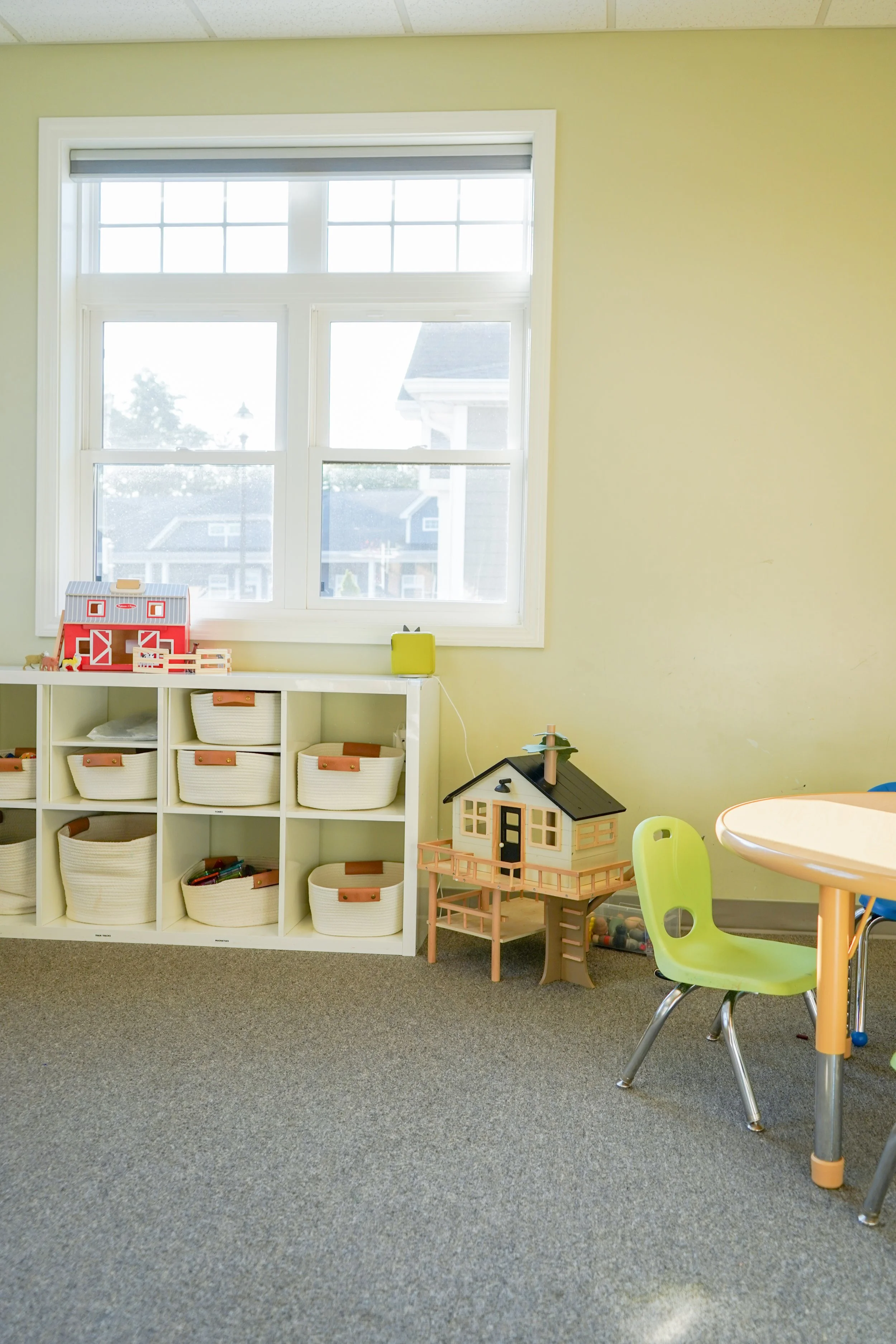 Child's playroom with a large window, green walls, and a gray carpet. Features a white storage unit with baskets, a dollhouse, and a small table with a yellow chair.