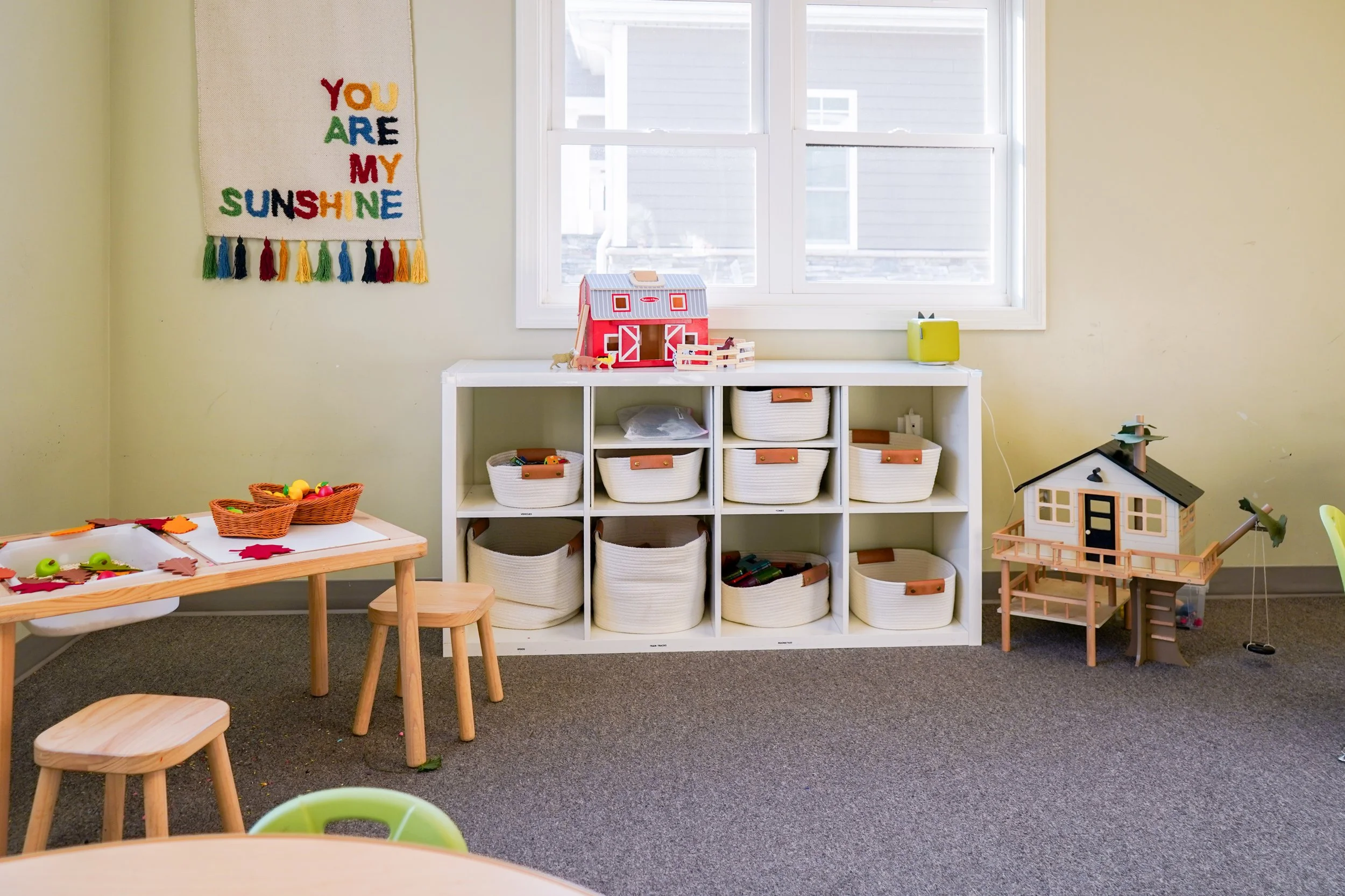 A children's playroom with a white cube storage unit holding baskets, a small table with chairs, and various toy houses and figures, decorated with a colorful wall hanging that says 'You Are My Sunshine' and a window letting in natural light.