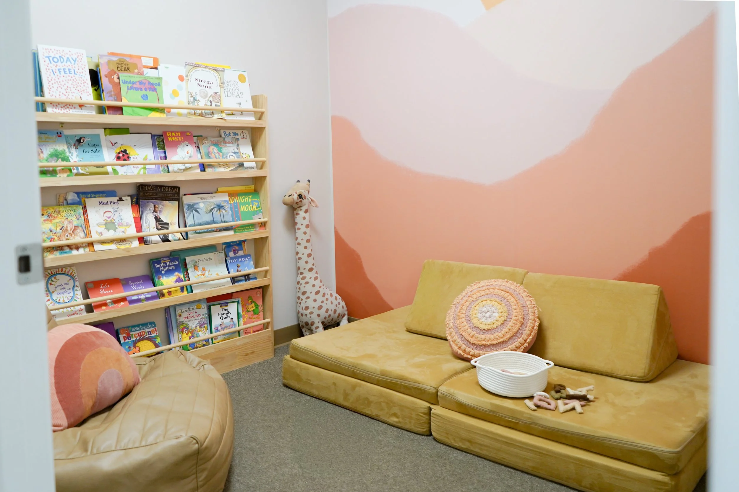 A cozy children's reading nook with a yellow couch, a pink and orange circular pillow, a white basket, and wall decorations. A bookshelf filled with children's books is on the left, and a plush giraffe toy stands in the corner by a pink and beige abs