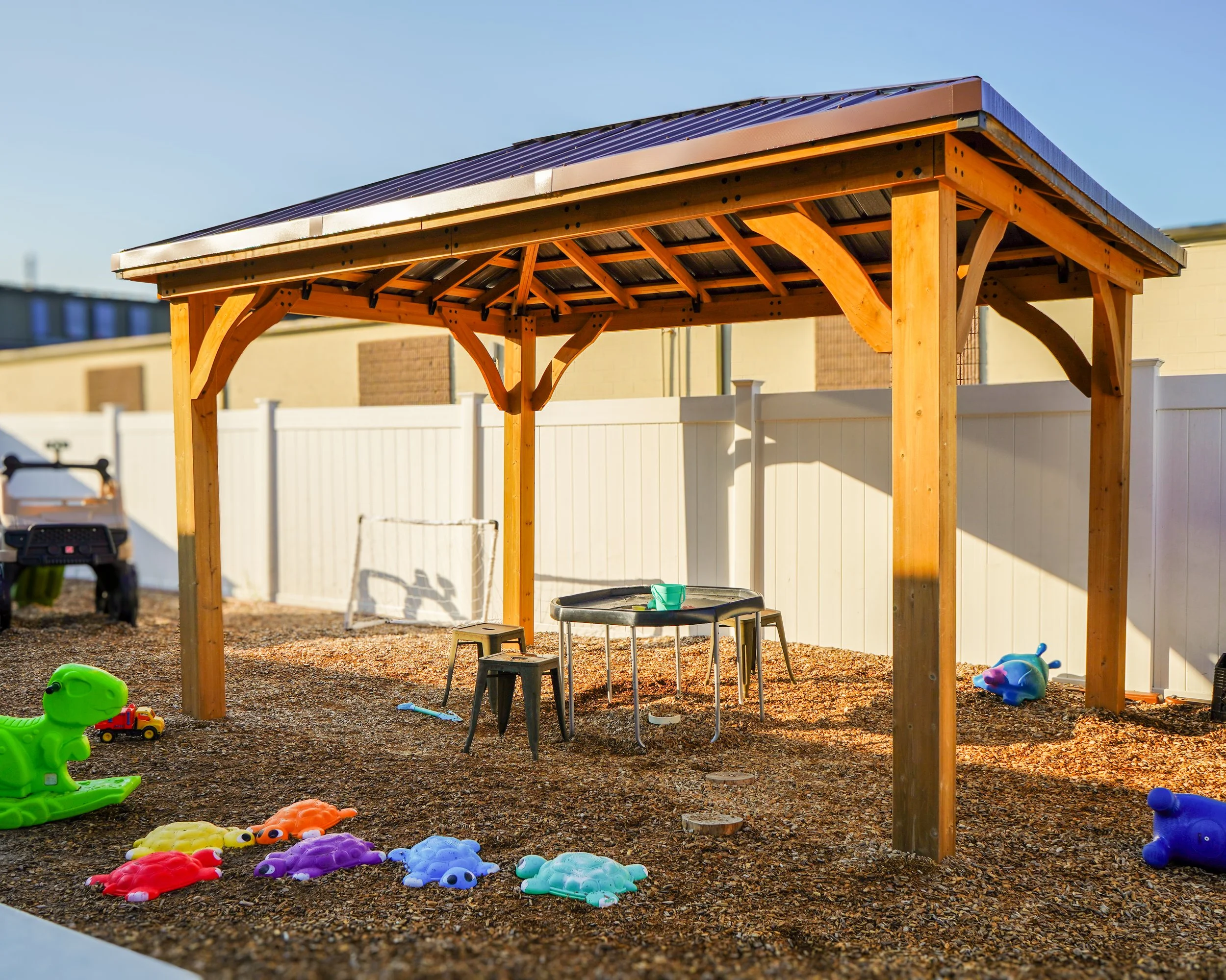 Backyard play area with a wooden canopy, plastic toys including dinosaurs, a small table with chairs, and a fenced yard.