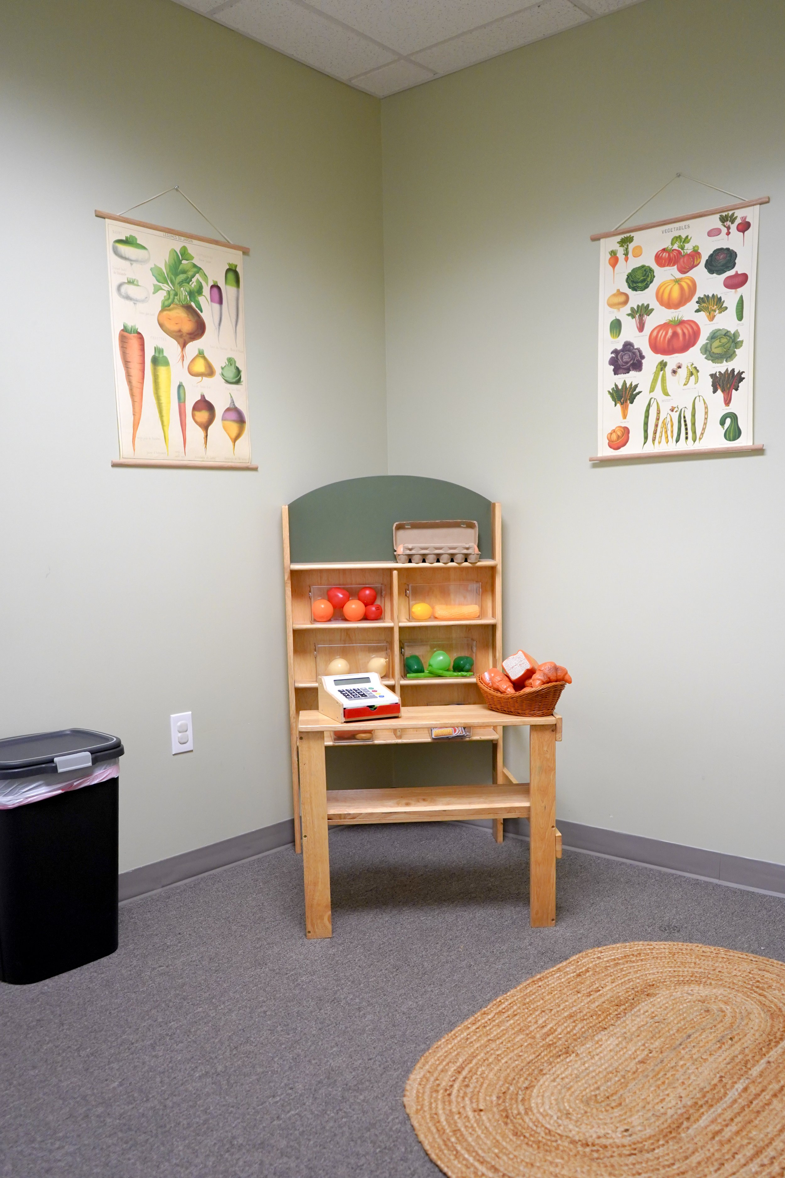 Play area with wooden supermarket cash register, toy vegetables, and posters of fruits and vegetables on the wall.
