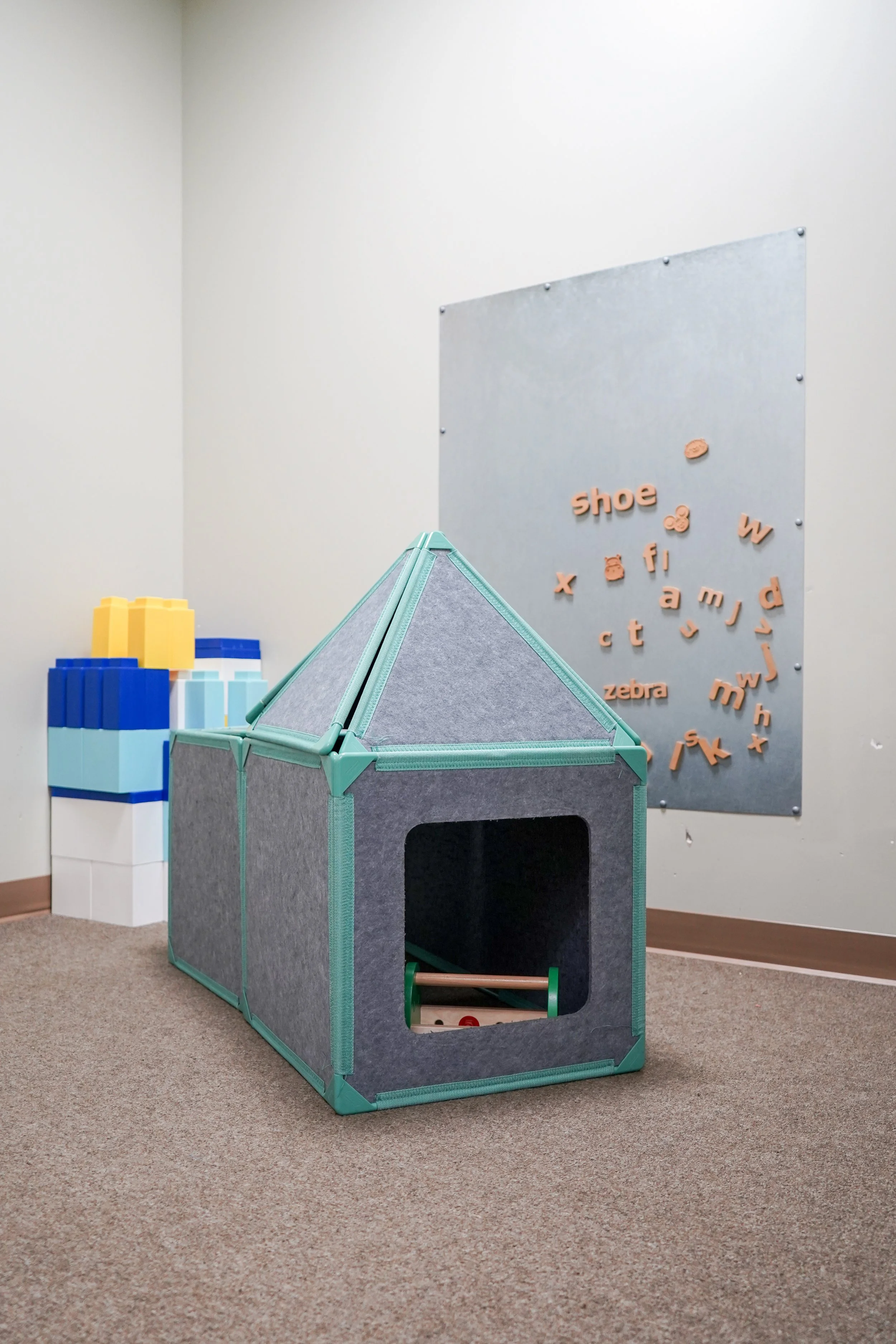 Children's play area with a grey fabric playhouse with teal trim, and foam blocks in yellow, blue, light blue, and white in the background, alongside a wall with letter magnets.
