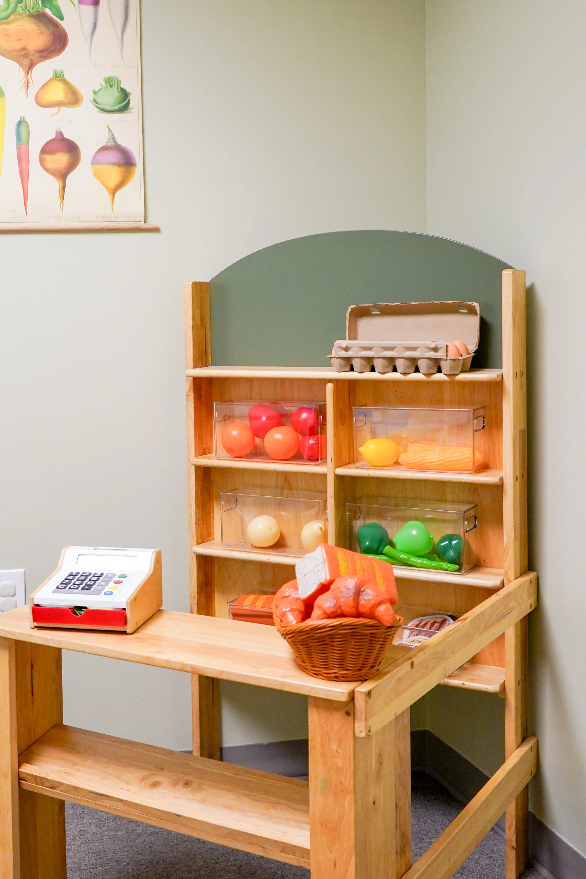 A wooden play grocery store with plastic produce and eggs, a basket of bread, and a toy cash register in a children's playroom.