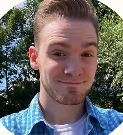 Close-up of a young man with light brown hair, wearing a blue plaid shirt, outdoors with trees and blue sky in the background.