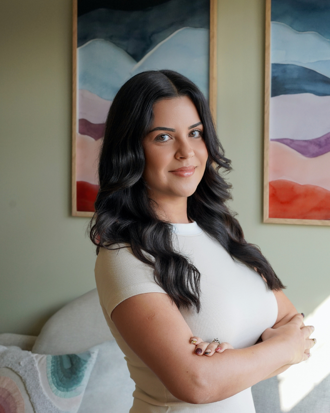 A woman with long dark wavy hair standing with her arms crossed in a room decorated with abstract landscape paintings on the wall behind her.