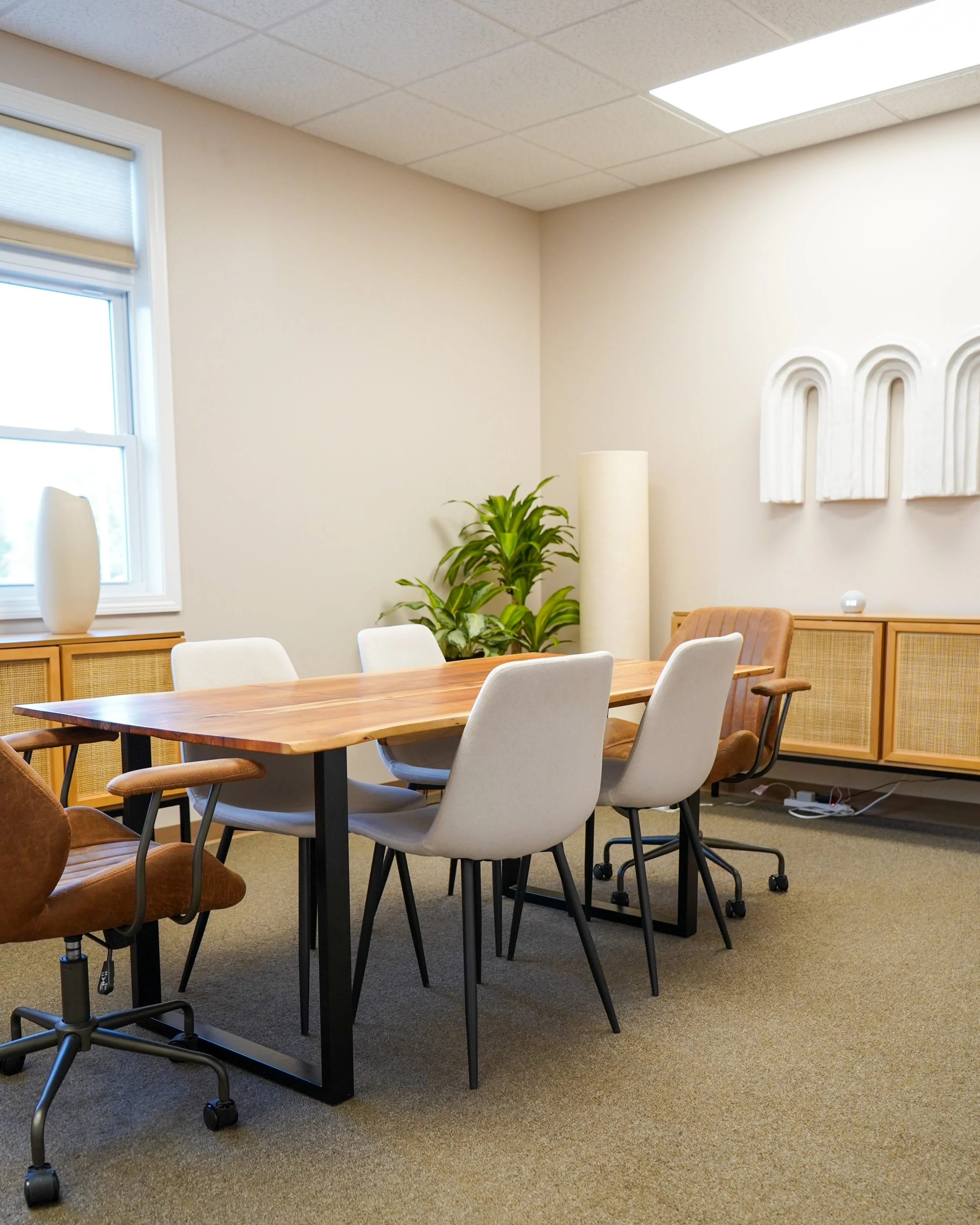 A meeting room with a wooden table, six chairs around it, some with beige upholstery and some with brown leather, a window with a blind, a sideboard with a white vase on top, a green potted plant, a tall white cylindrical lamp, and a wall decoration 