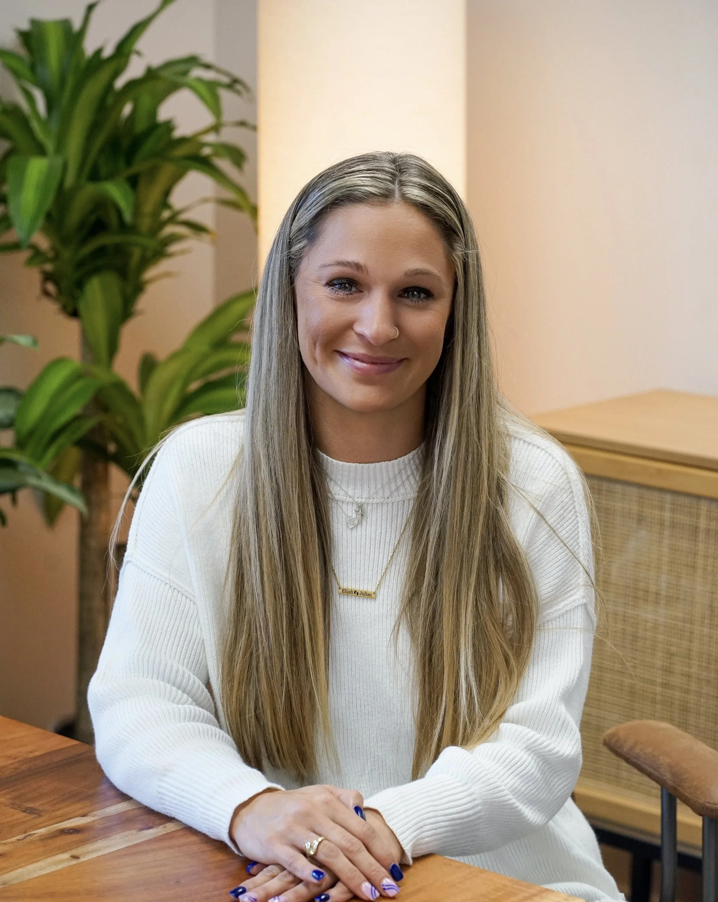 A woman with long blonde hair, wearing a white sweater, and smiling, sitting at a wooden table with a potted plant and a light-colored wall in the background.