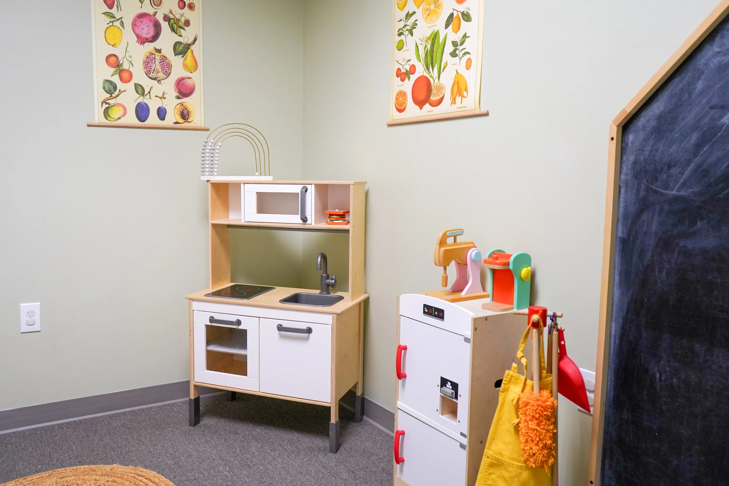 Children's play kitchen with a stove, sink, and oven, along with a toy cash register, set up in a playroom with botanical fruit and vegetable posters on the wall.