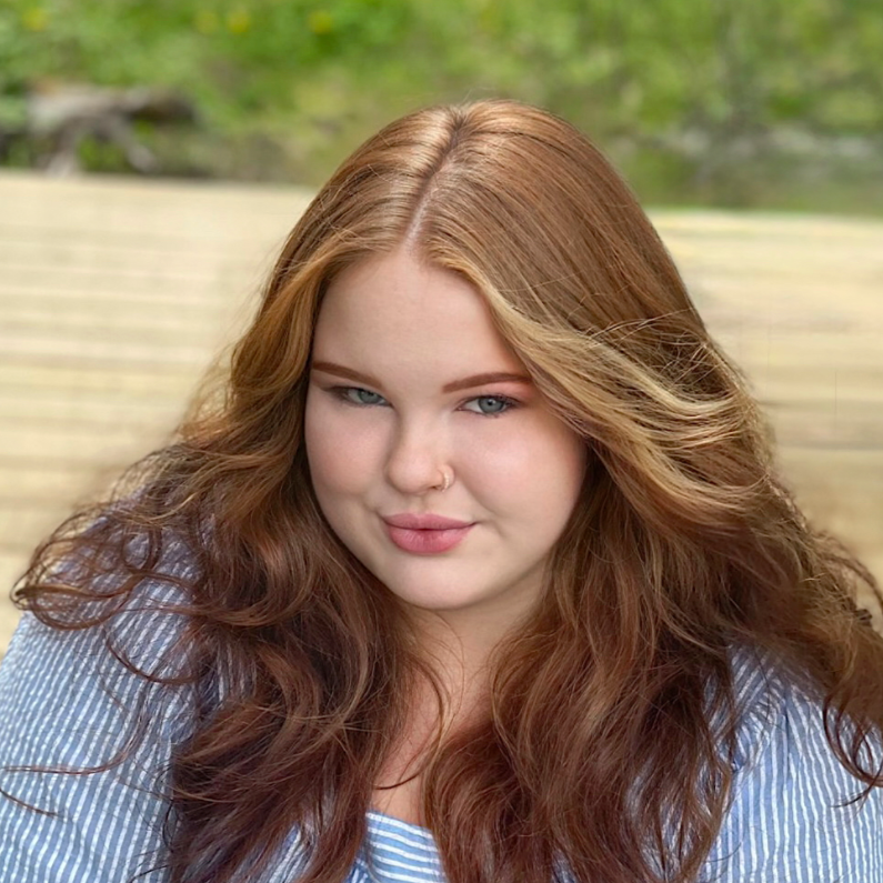 A young woman with long, wavy red hair and blue eyes. She has a nose piercing and is wearing a blue and white striped shirt. She is outdoors with blurred greenery in the background.
