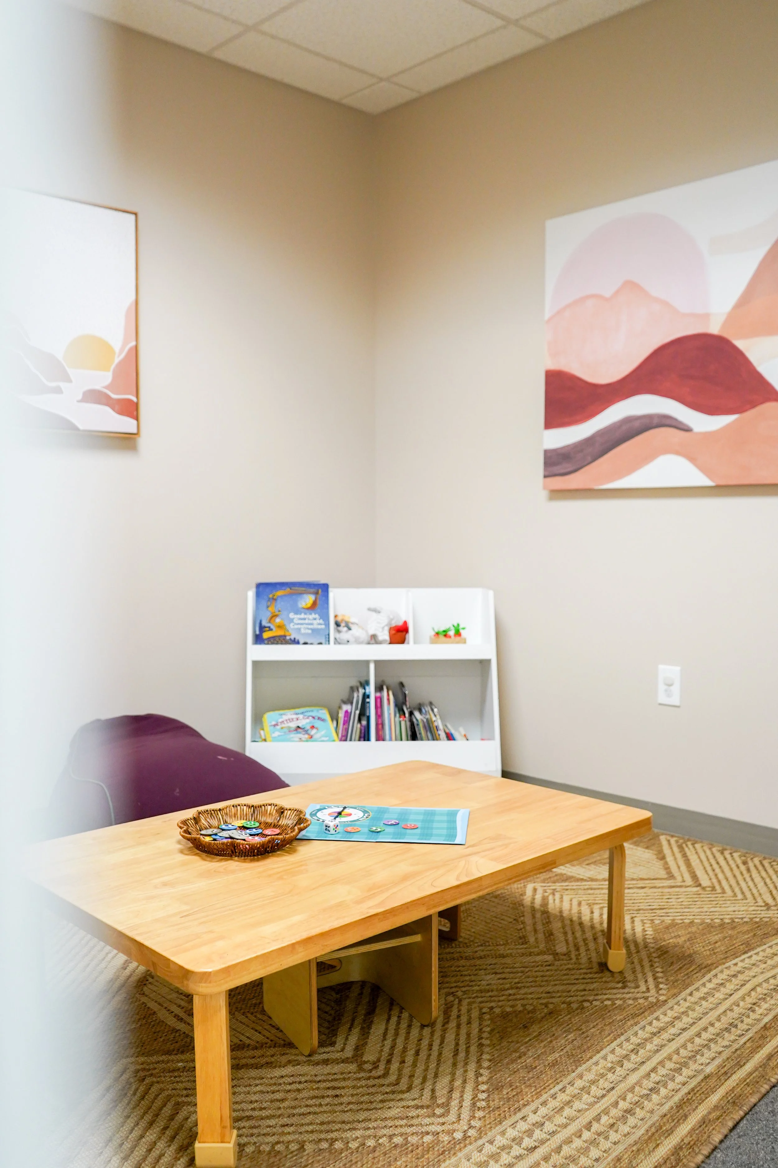 A small reading and play area with a low wooden table, a white bookcase filled with children's books and toys, and two colorful abstract landscape paintings on the beige walls.