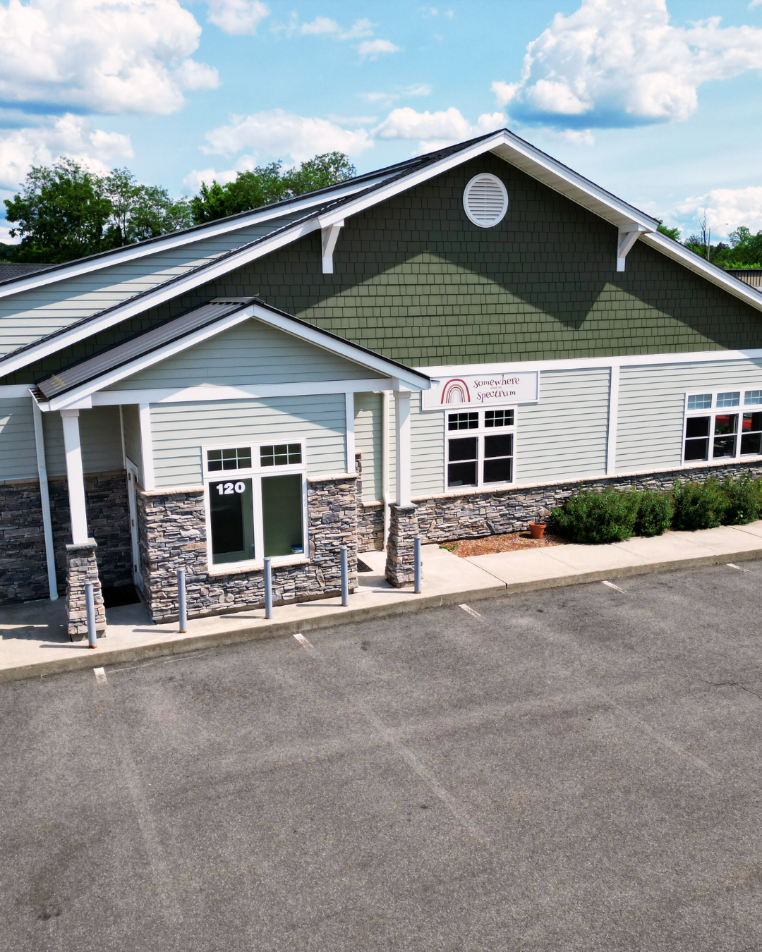 An exterior view of a modern commercial building with gray and green siding, double glass door entrance, and a sign that reads 'Somewhere Spectrum.' The building has stone accents at the base, surrounded by a parking lot and some landscaping with bus