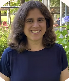 Woman with shoulder-length brown hair smiling outdoors, wearing a navy blue shirt, with greenery and flowers in the background.