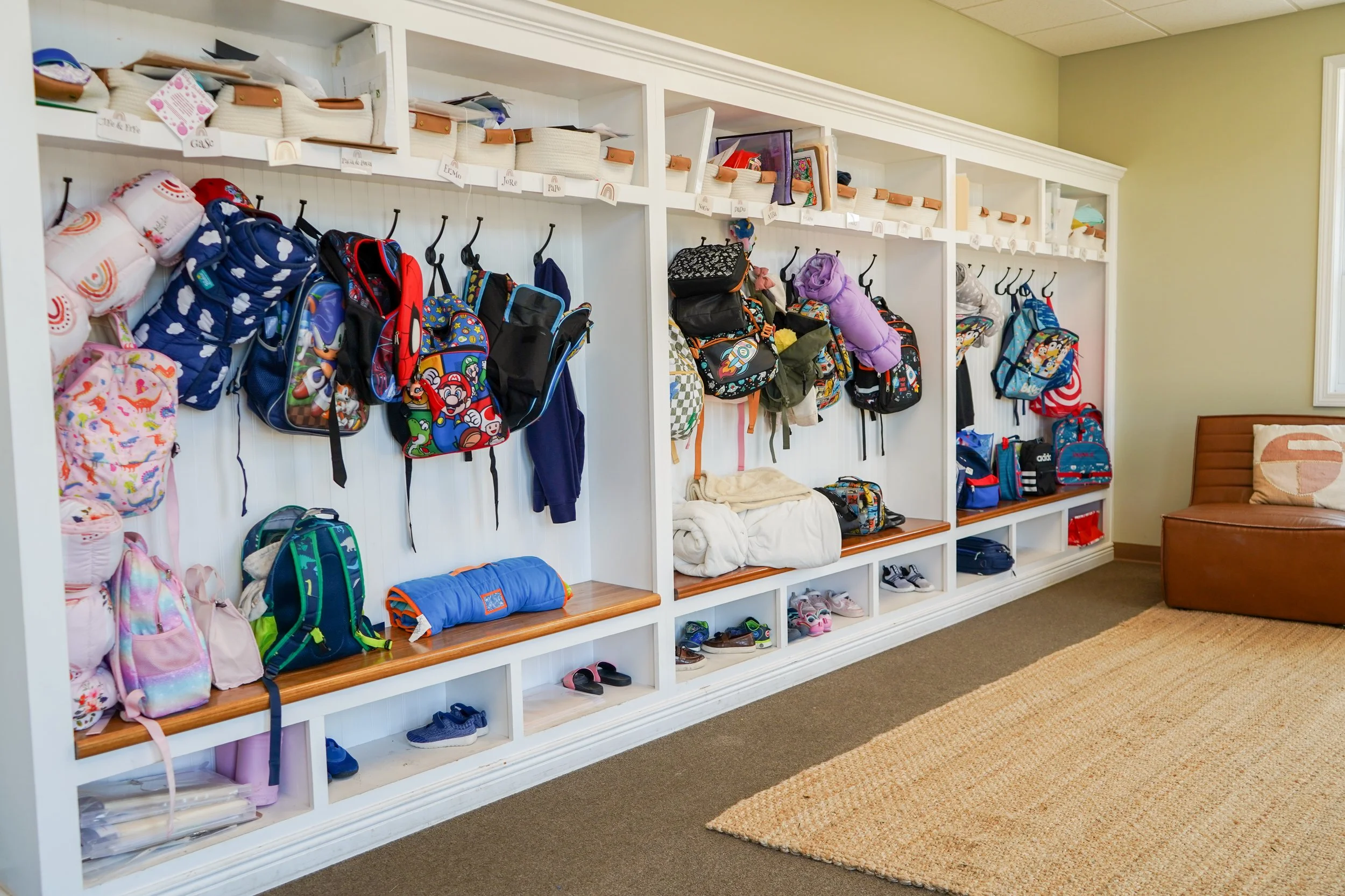 School hallway with white cubbies filled with backpacks, school supplies, and jackets, and a brown leather bench near the wall.
