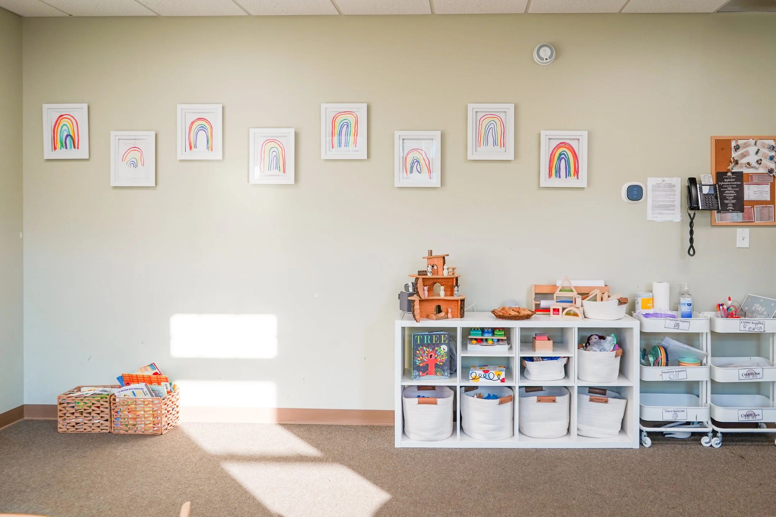 Child-friendly classroom corner with rainbow drawings on the wall, toy storage baskets, a dollhouse, and educational materials.