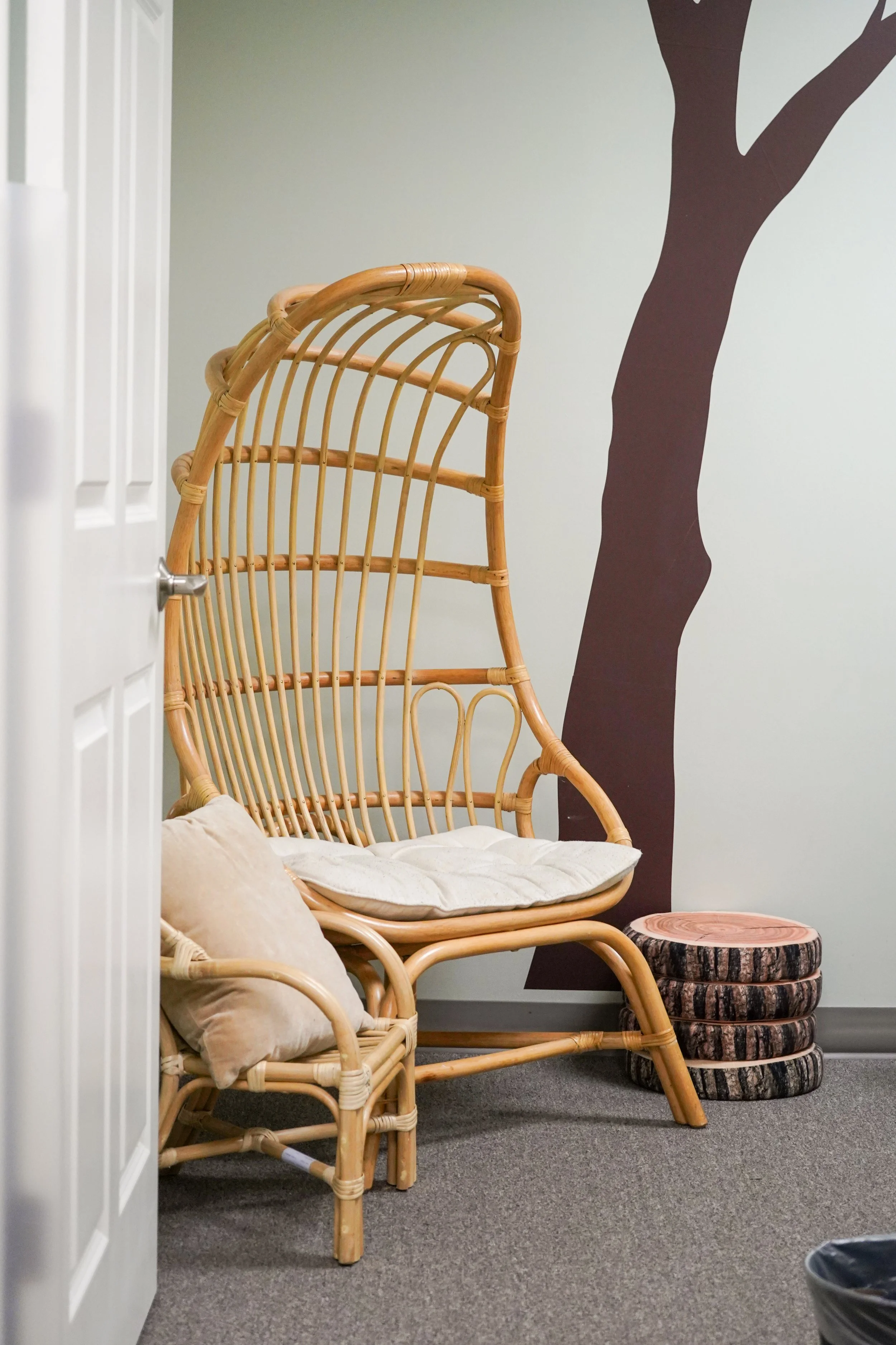 A rattan high-back chair with a cream cushion and a beige pillow, next to a small stack of round wooden logs, in front of a wall with a dark brown tree silhouette decal.