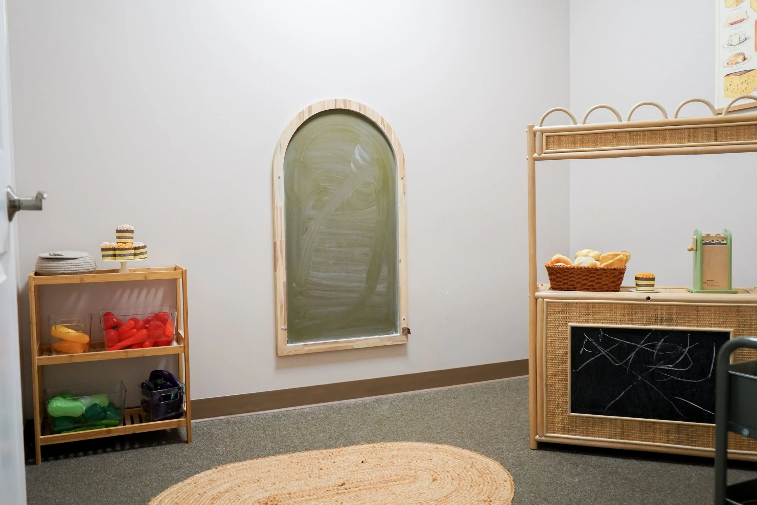 Empty retail or play area with shelves holding plastic food and toys, chalkboard, and chalk, with a oval rug on carpeted floor, and a window with faux window dressing.