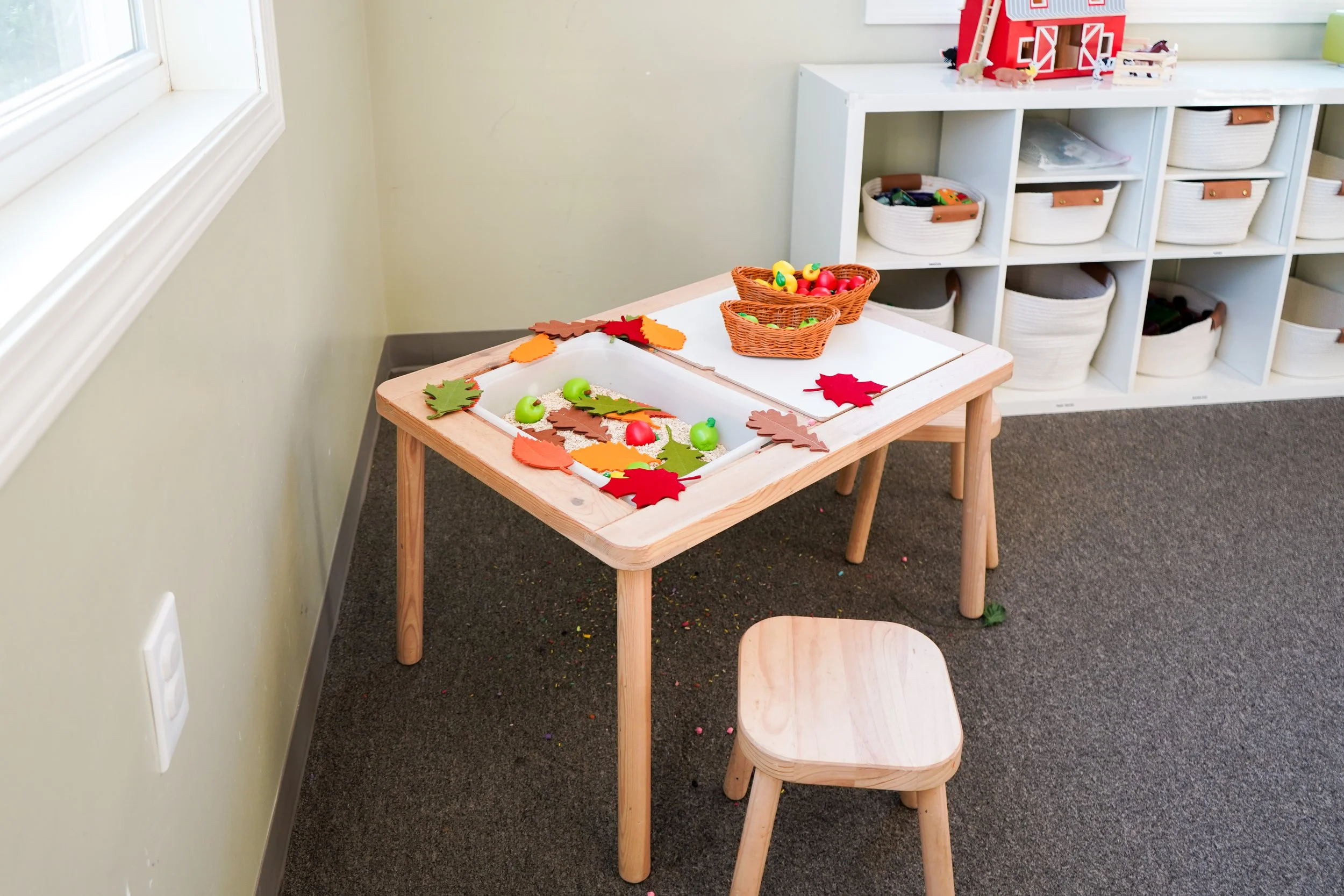 A children's play area with a small wooden table and stool. The table is decorated with autumn leaves and small toys, with a basket of plastic eggs on top. In the background, there is a white shelf with storage baskets and a toy barn on top.